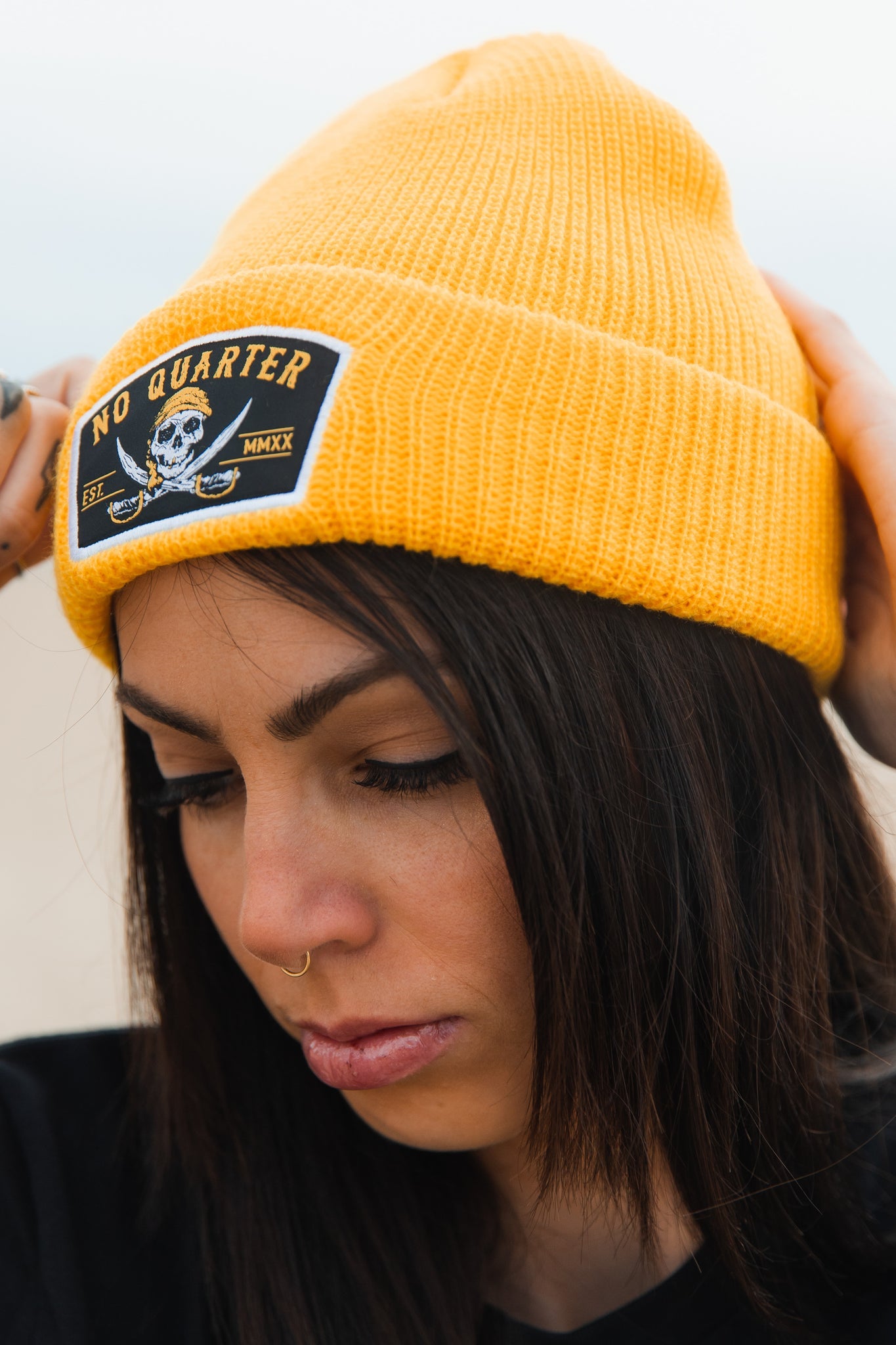 A woman with long, dark hair and a nose ring wears a yellow No Quarter Beanie by Relentless Betrayal, featuring a skull and crossbones. Her expression is neutral as she looks downward against a blurred background.