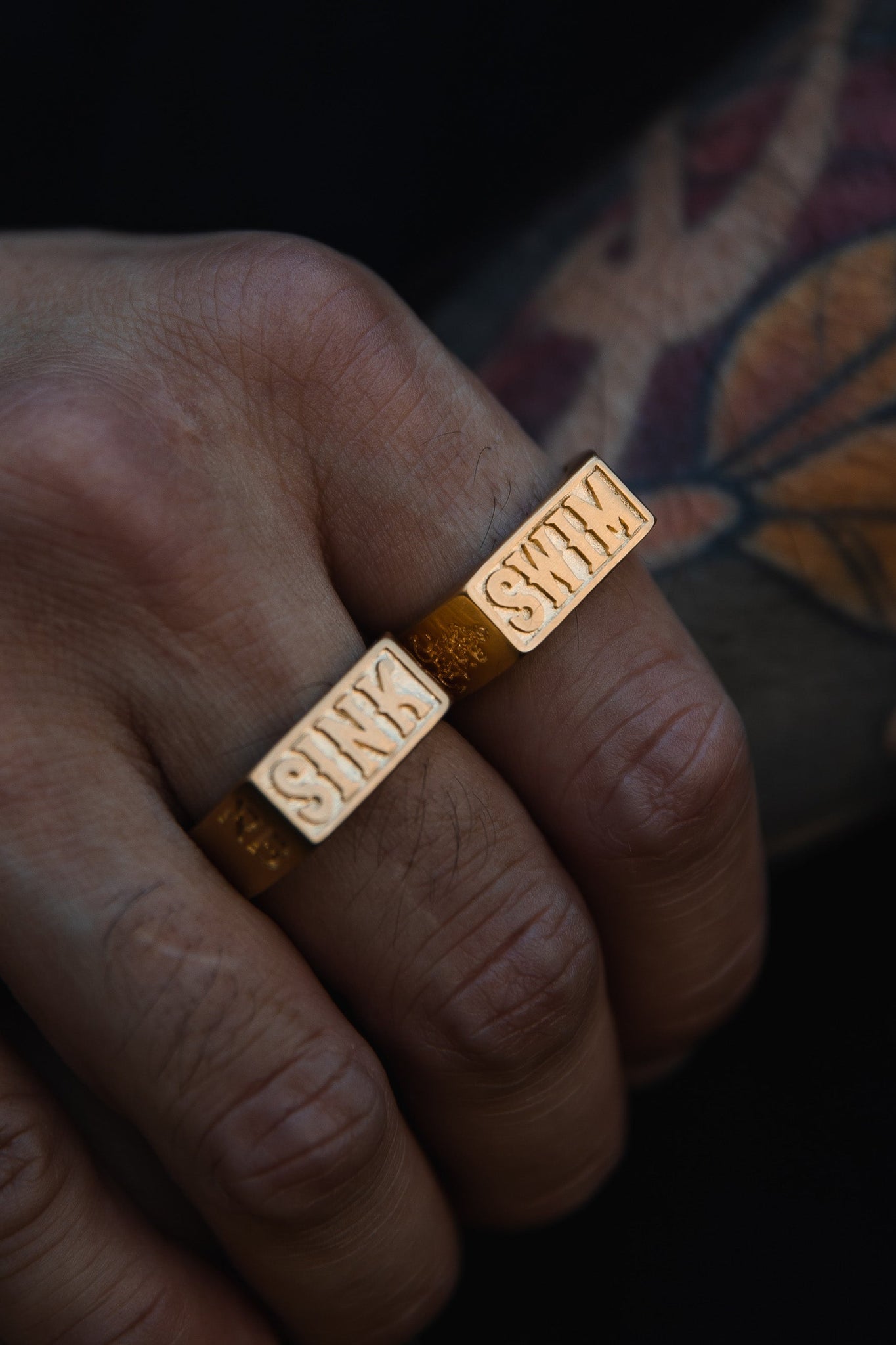 A hand wearing two Relentless Betrayals Gold Sink or Swim Rings, each engraved with SINK, rests side by side. The bold rings are complemented by a colorful tattoo on the skin, showcasing a unique blend of art and style.