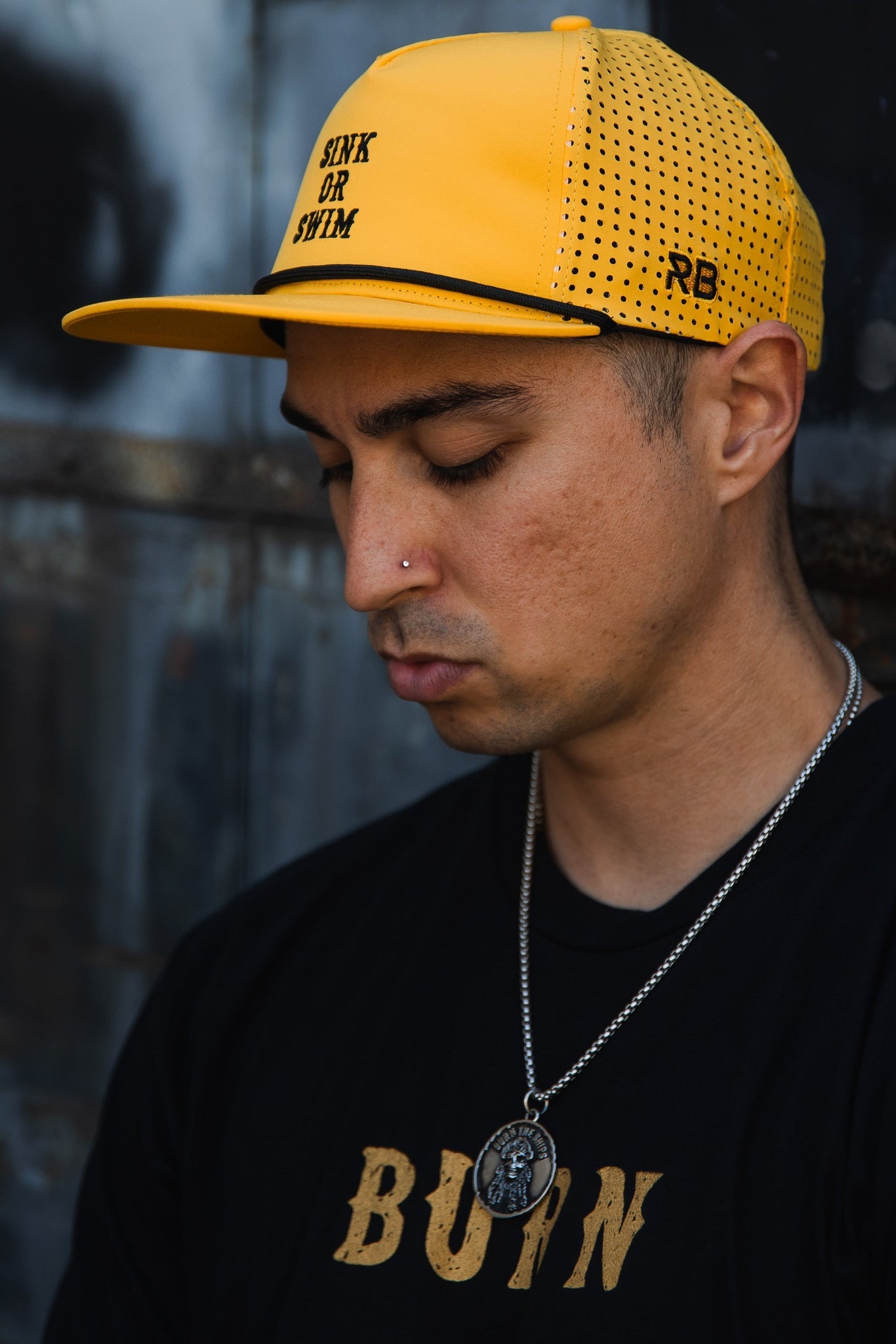 A person wearing a handmade yellow Gold Sink or Swim Snapback by Relentless Betrayal and a black shirt with BUIN in gold letters. They have a nose piercing, gaze down, and wear a necklace against a blurred wall backdrop.