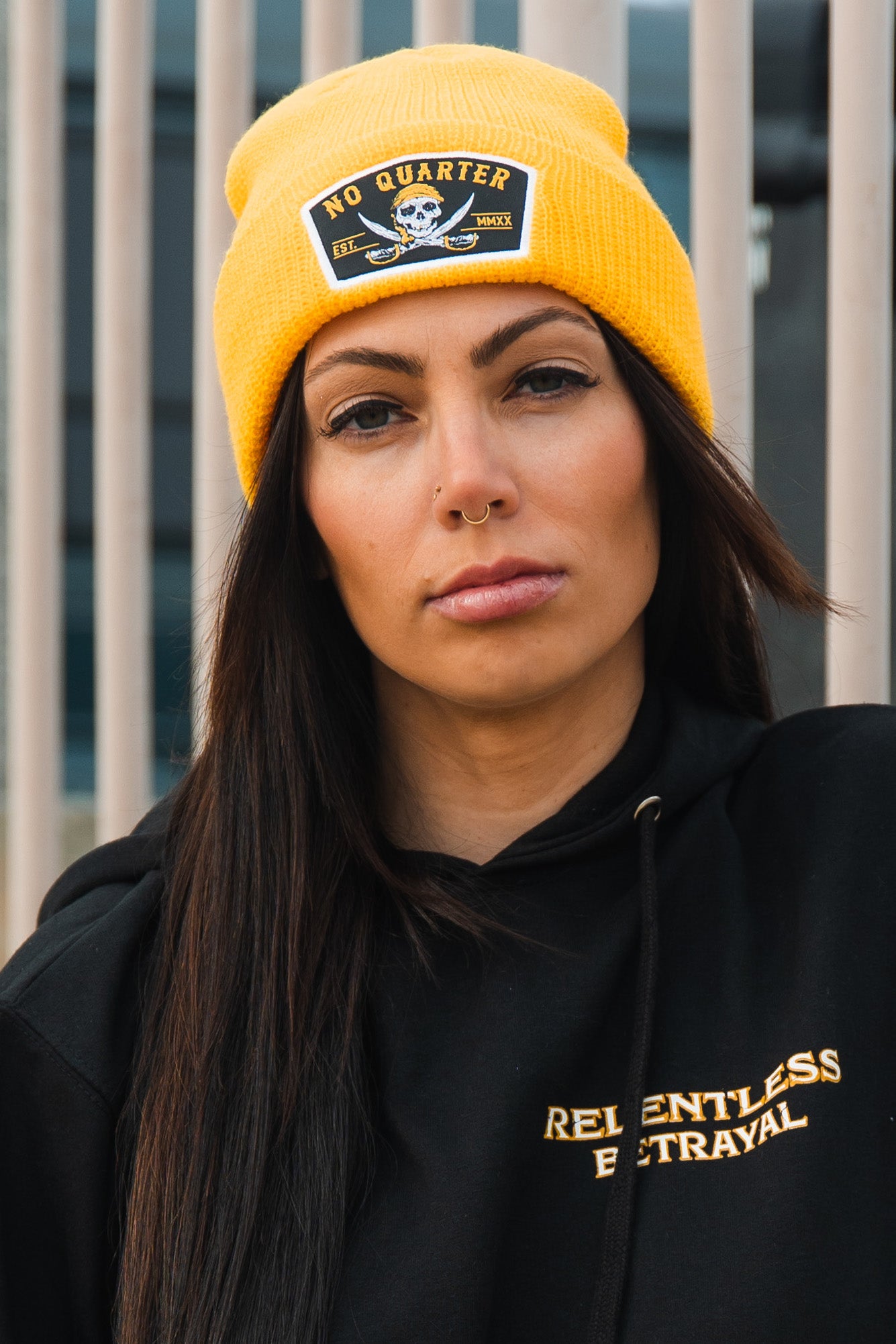 A person confidently stands in front of a metal fence, wearing the No Quarter beanie from Relentless Betrayal along with a black hoodie featuring the brands name.