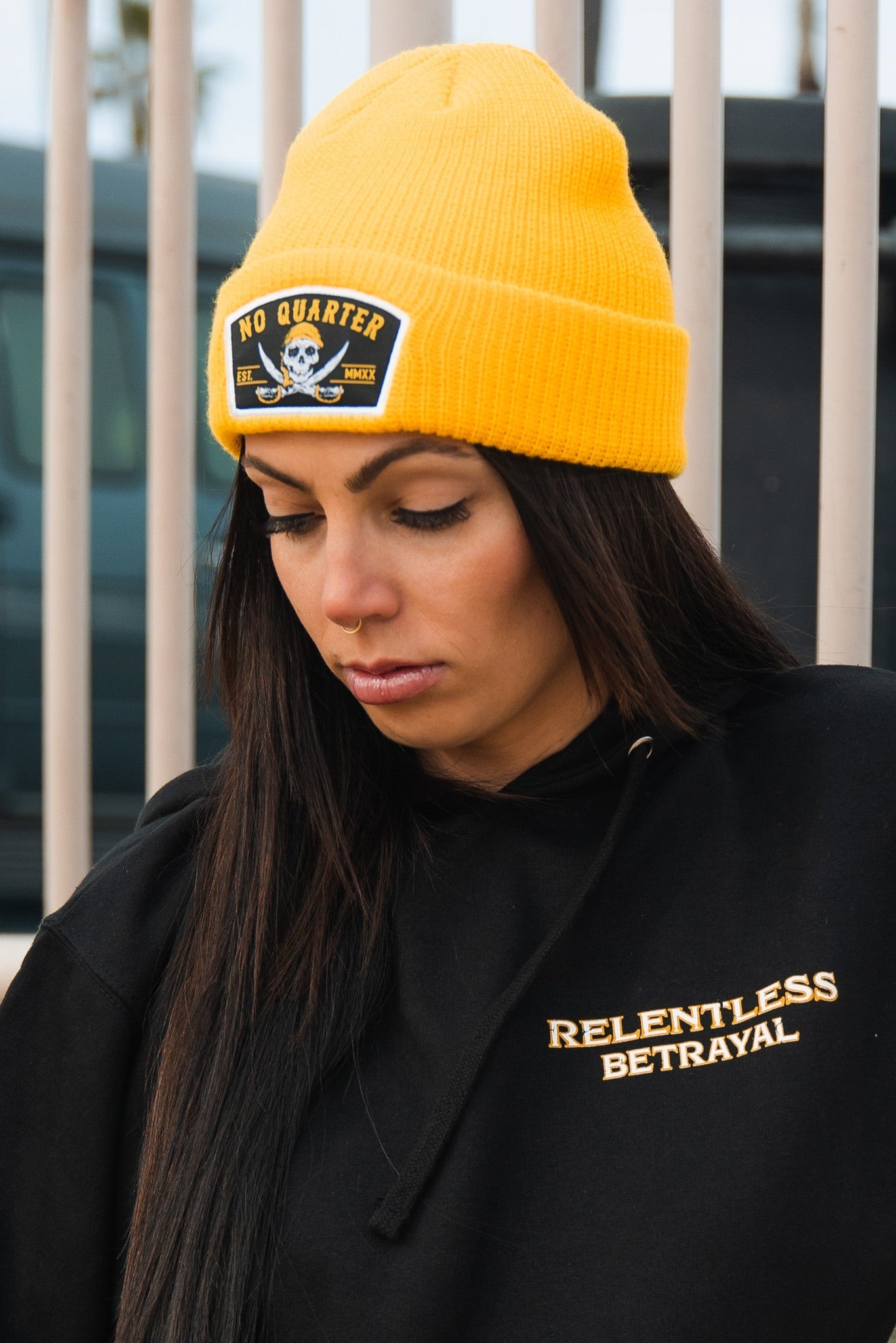 A person with long dark hair wears a bright yellow No Quarter Beanie, featuring a skull and crossbones patch, along with a black hoodie from Relentless Betrayal. A fence and vehicle are seen behind them.