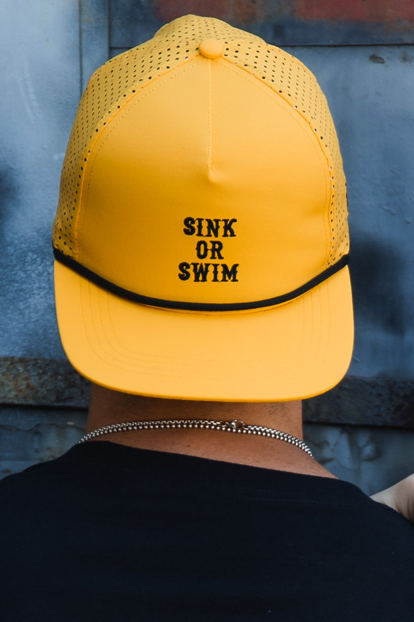 A person wears a handmade Gold Sink or Swim Snapback by Relentless Betrayal, featuring a bright yellow front with SINK OR SWIM in bold black letters, mesh back, and curved brim. Seen from behind in a black shirt.