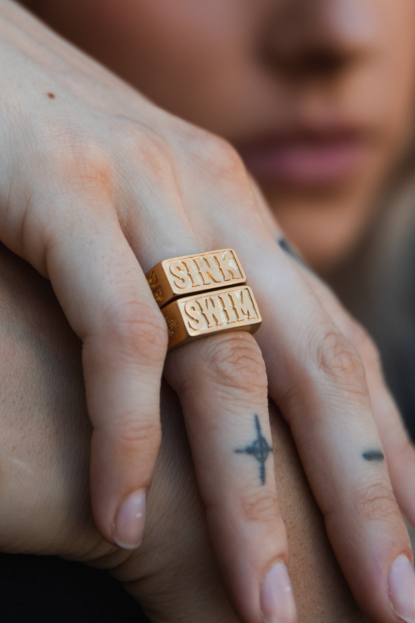 A close-up shows a hand resting on another, wearing two Relentless Betrayals handcrafted Gold Sink or Swim Rings. The fingers feature simple tattoos, like a cross on the middle finger, with a softly blurred background enhancing this intimate scene.