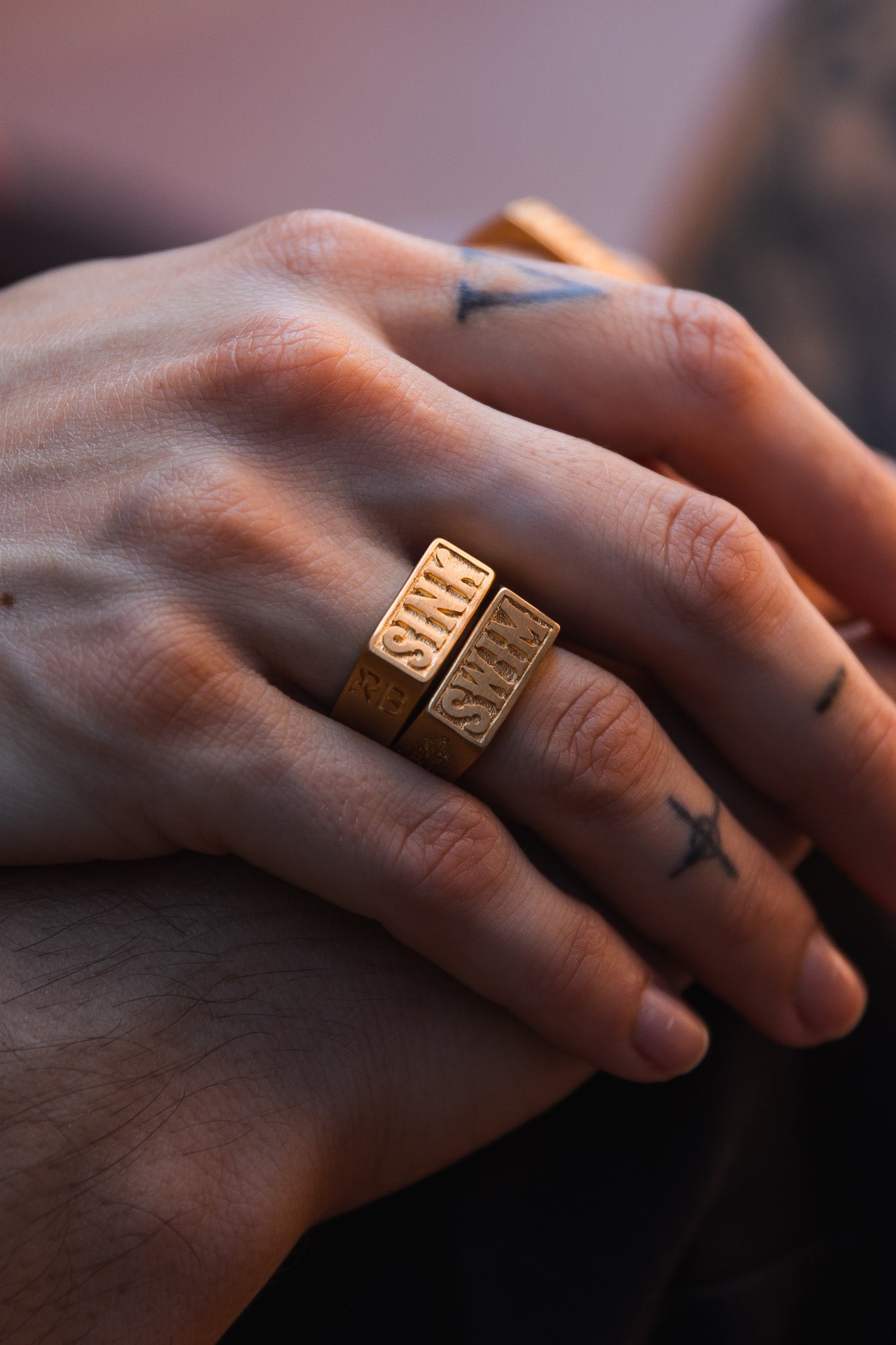 Close-up of hands wearing two Gold Sink or Swim Rings by Relentless Betrayal, inscribed with SMILE, and a handcrafted stainless steel ring. Small tattoos like a cross and triangle adorn the fingers, resting gently above another hand in soft, warm lighting.