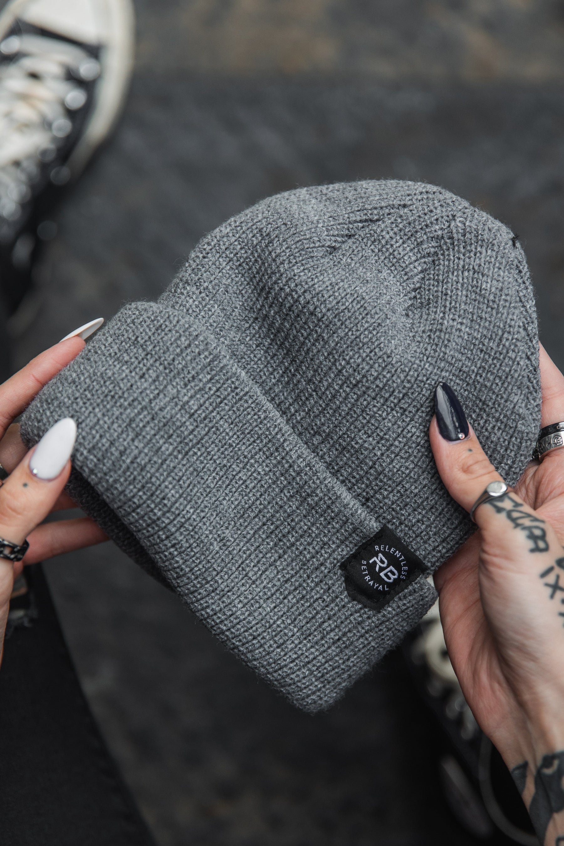 Tattooed hands with rings hold a Relentless Betrayal Beanie featuring a black logo patch. The individuals long black and white nails complement the sneakers in the background on a dark surface, enhancing the casual style of this unisex accessory ensemble.