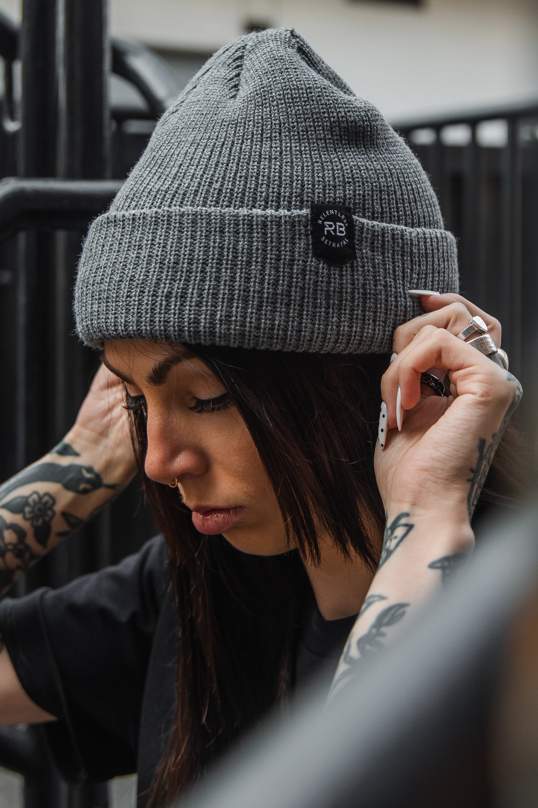 A person with tattoos wearing a Relentless Betrayal Beanie and a black shirt is adjusting their earphones. They have long dark hair and stand near black metal bars, with the focus on their face and hands.