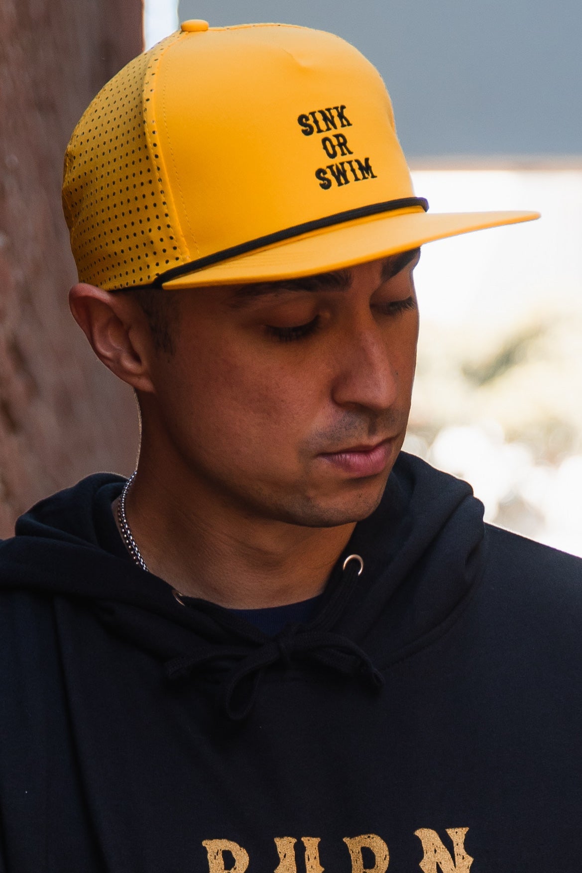 A person in a black hoodie wears a yellow, water-resistant Gold Sink or Swim Snapback from Relentless Betrayal, with their head down against a blurred background.
