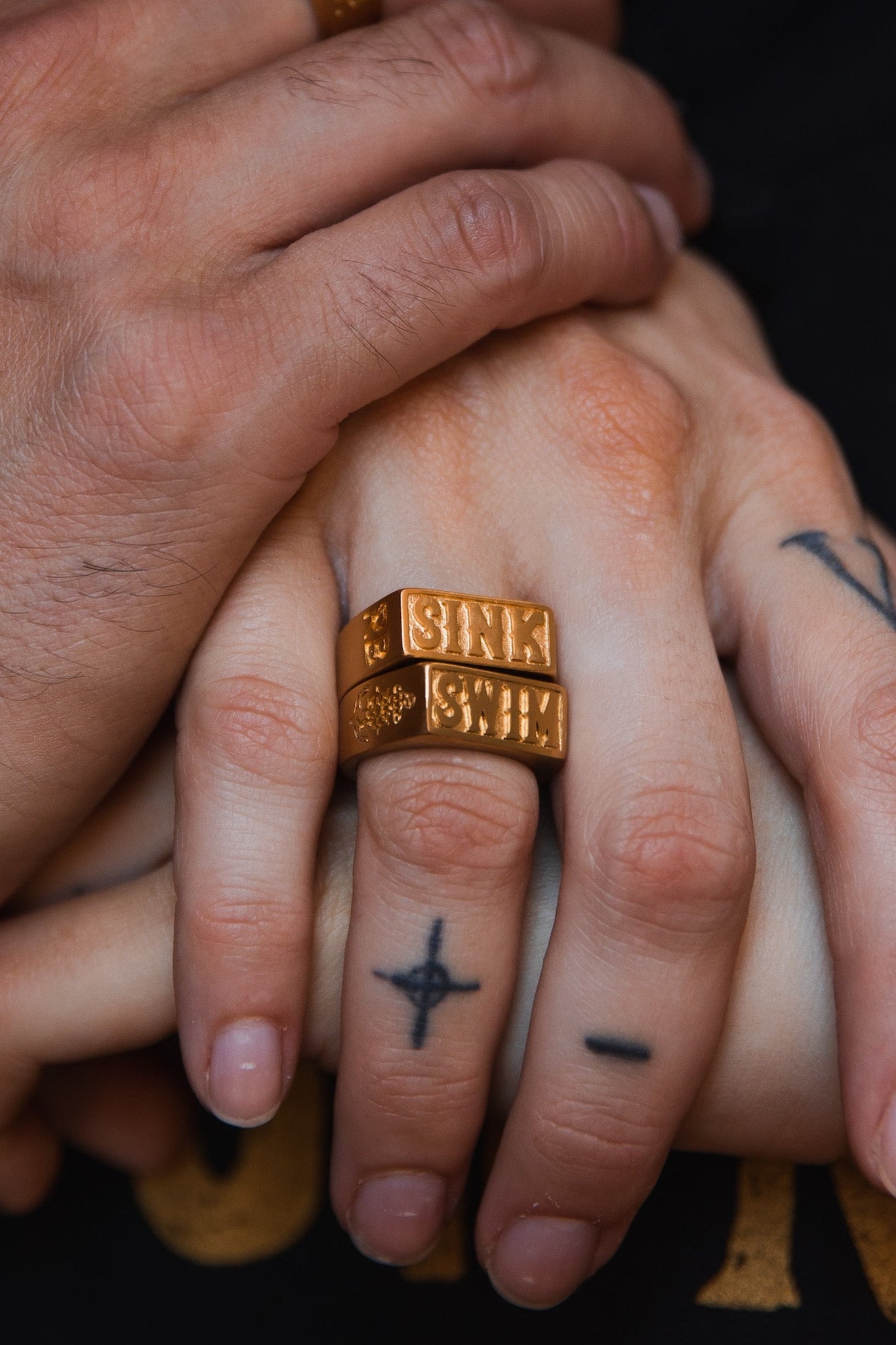 Close-up of tattooed hands wearing handcrafted Gold Sink or Swim Rings by Relentless Betrayal, entwined. The rings, engraved with block letters, rest among fingers inked with small crosses and lines, on skin tones ranging from light to medium.