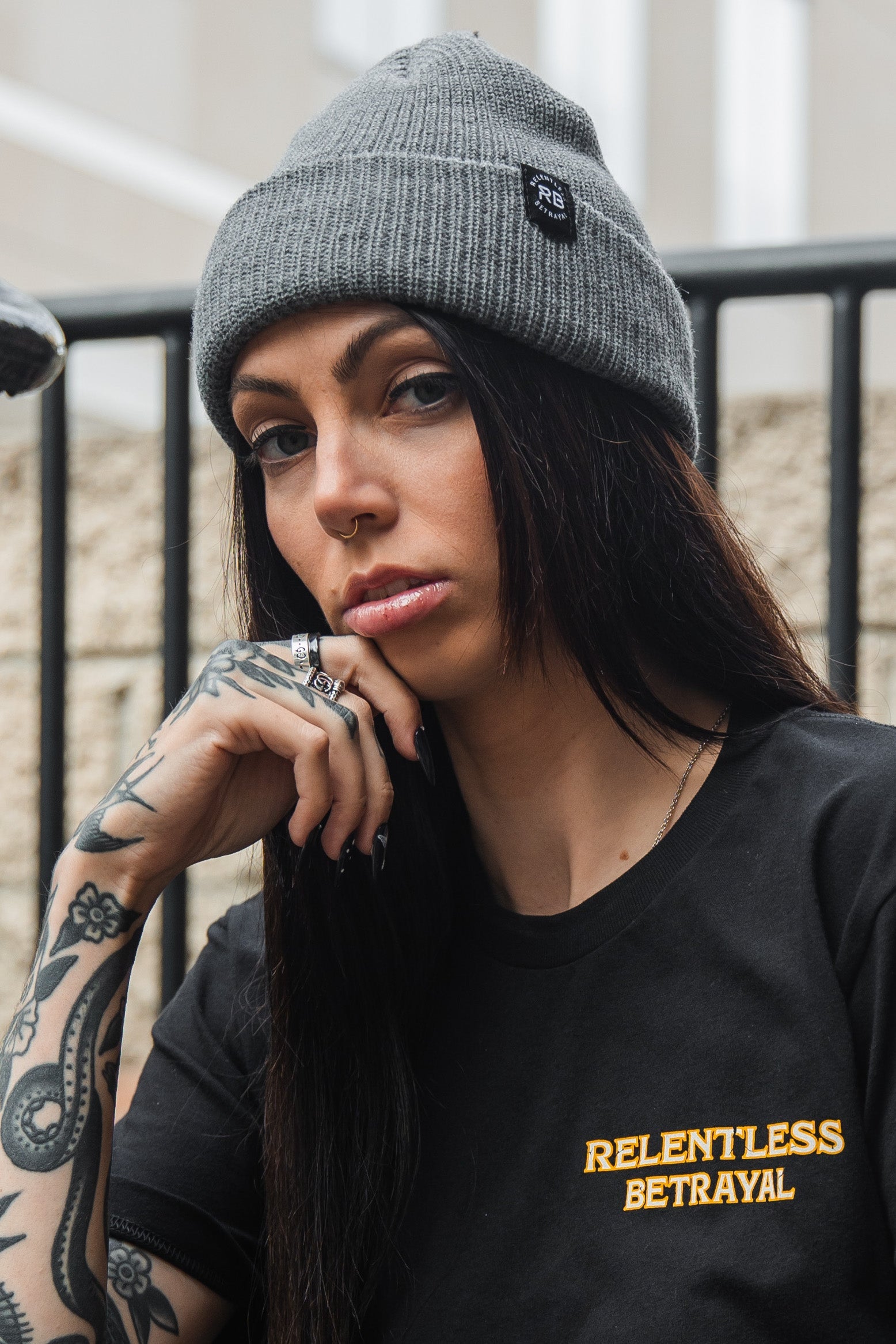 A person with long dark hair and tattooed hands wears a gray knitted Relentless Betrayal Beanie and a black T-shirt reading RELENTLESS BETRAYAL, sitting outdoors, chin in hand, gazing directly at the camera.
