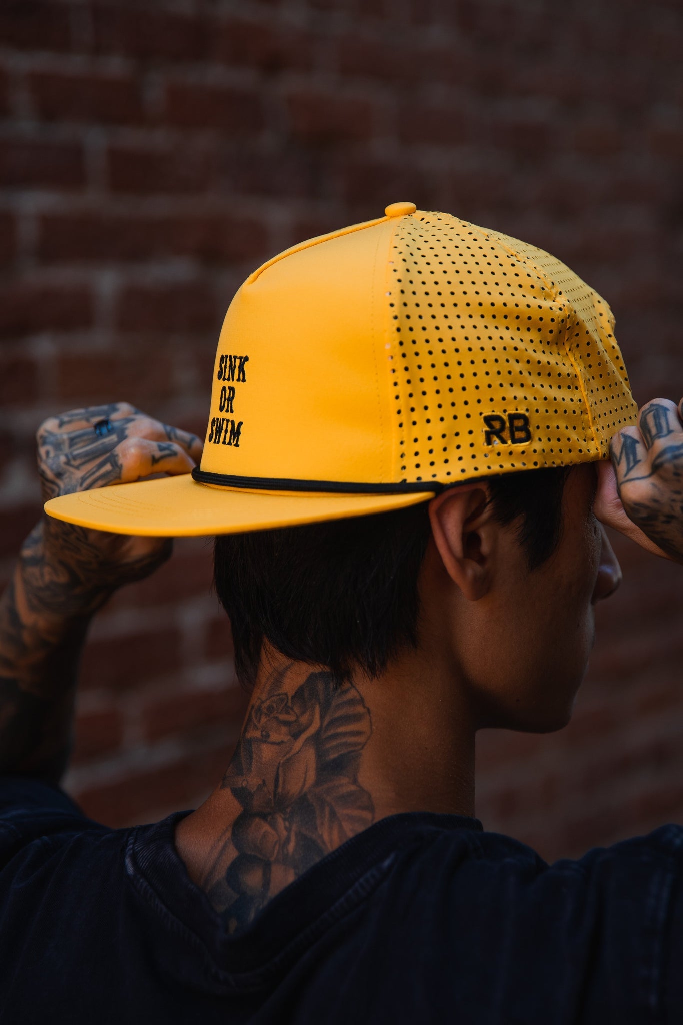 A person with tattooed arms and neck adjusts a handcrafted Gold Sink or Swim Snapback by Relentless Betrayal while facing a brick wall.