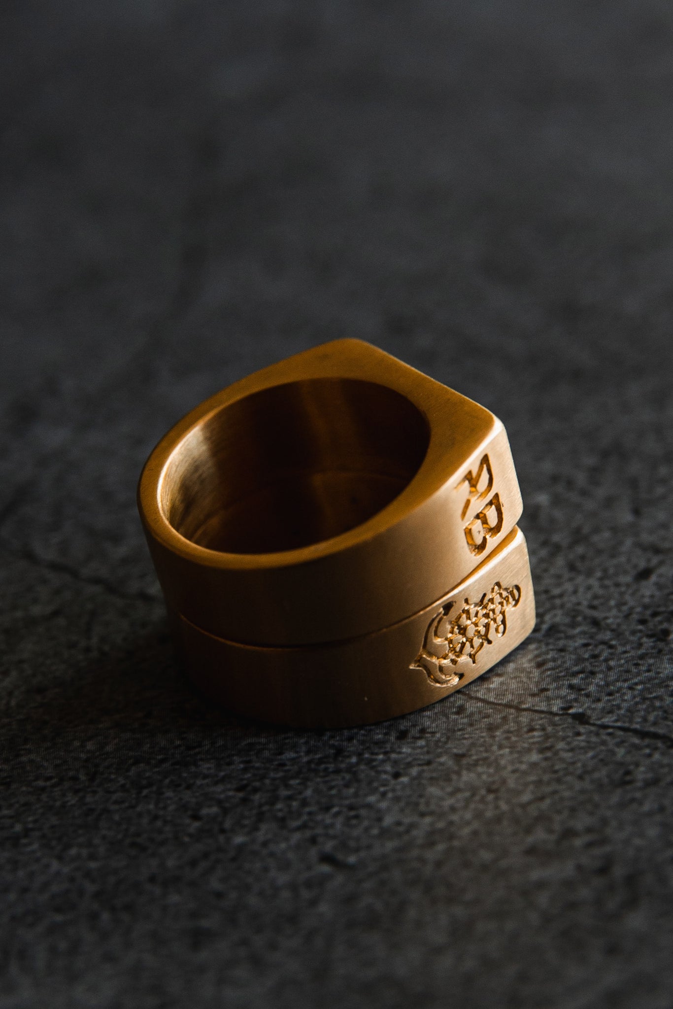 The Gold Sink or Swim Rings by Relentless Betrayal, one engraved with RB and the other featuring a lions head design, capture attention as they are stacked on a dark, textured surface. Their metallic sheen is highlighted beautifully by the lighting.