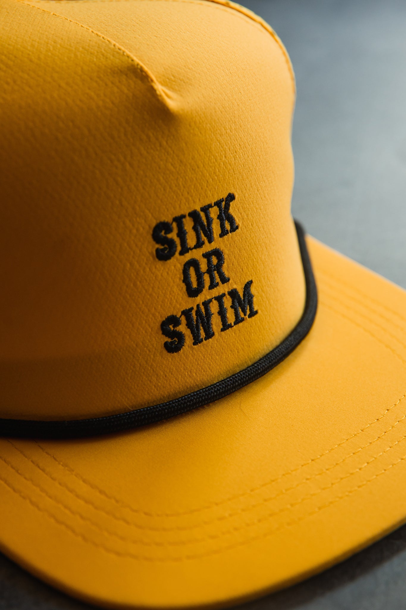 Close-up of the Gold Sink or Swim Snapback by Relentless Betrayal, showcasing black embroidered text SINK OR SWIM. The yellow cap rests on a dark surface with a black trim around the brim.