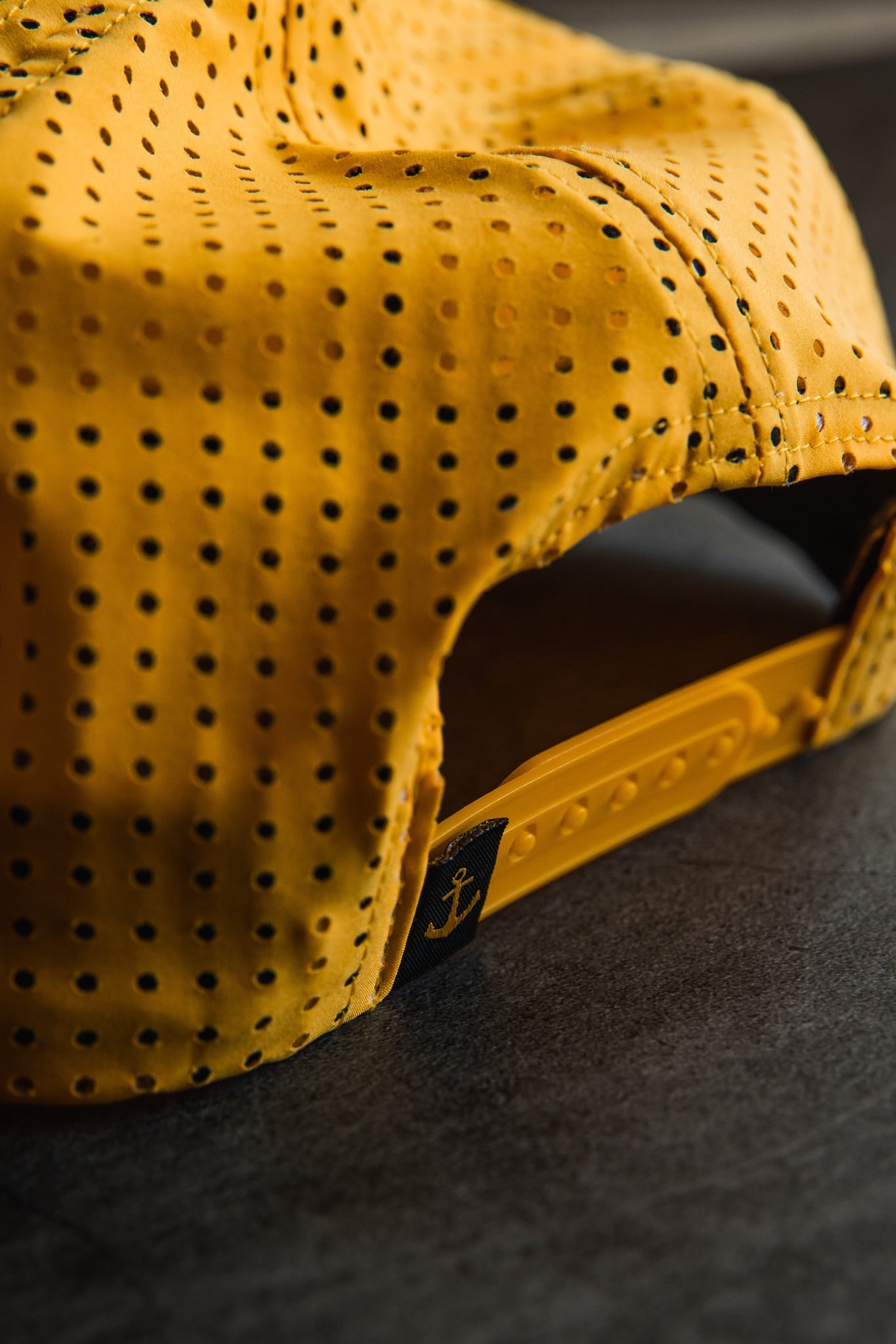The Gold Sink or Swim Snapback by Relentless Betrayal is beautifully highlighted with close-up details, featuring a yellow hue and plastic closure. An anchor symbol graces a small black tag near the clasp, accentuating its charm as it rests on a dark surface, showcasing unique style and craftsmanship.