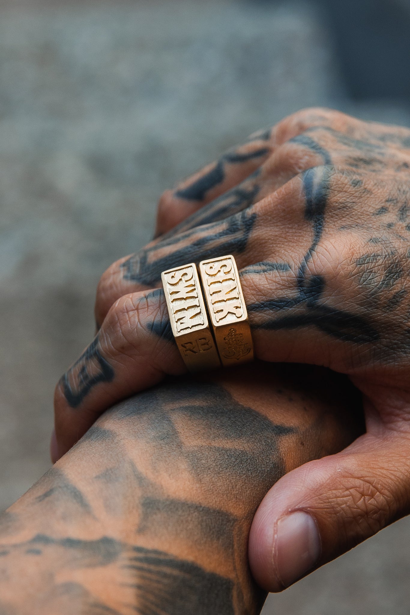 Close-up of tattooed hands, one holding the other. The hand displays two thick handcrafted Gold Sink or Swim Rings from Relentless Betrayal, with intricate SKTS engravings and tattoos featuring complex patterns against a softly blurred background.