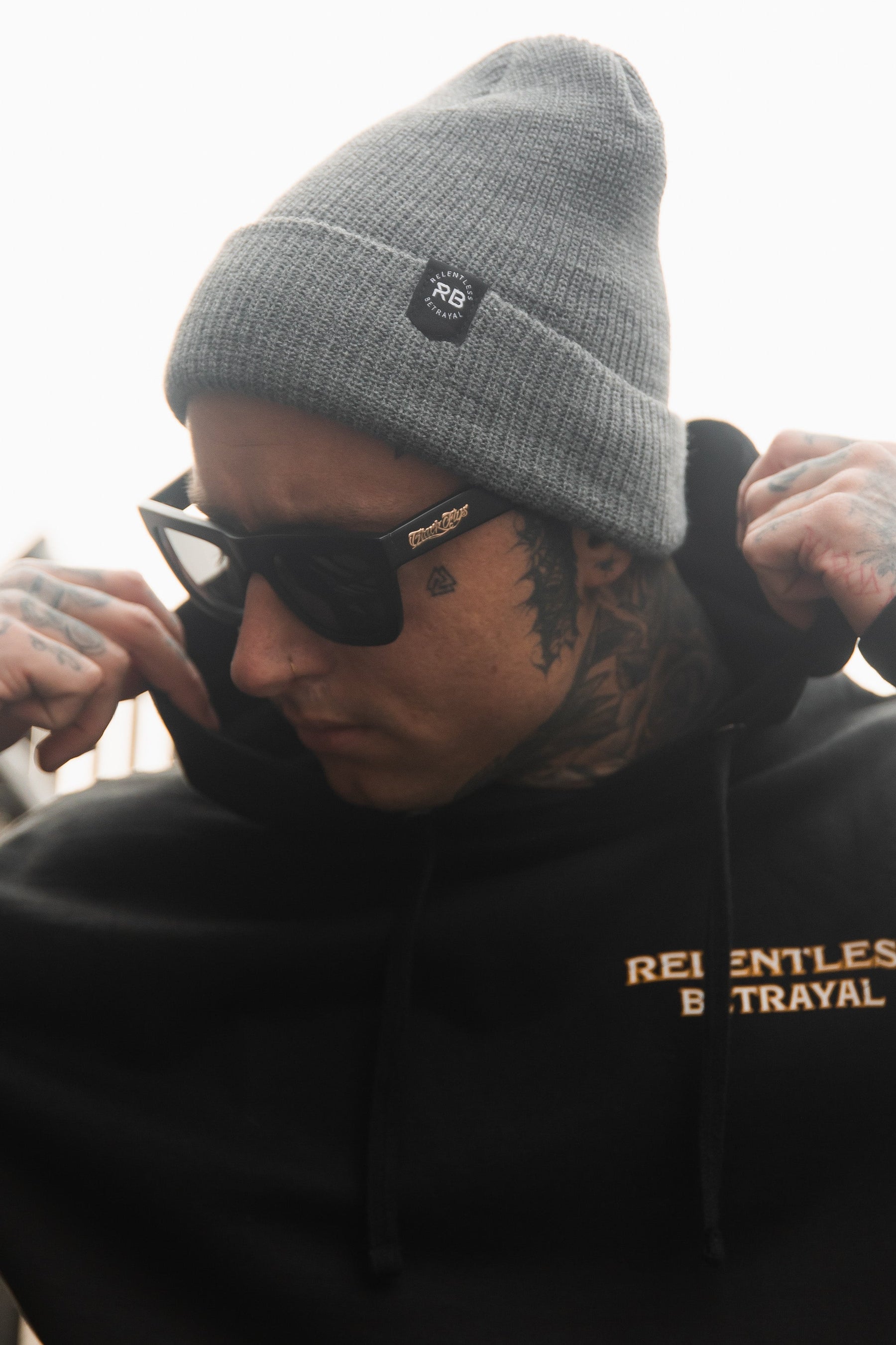 A person in a gray Relentless Betrayal Beanie and black sunglasses adjusts their black hoodie with Relentless Betrayal text, showcasing tattooed neck and hands for an edgy style.