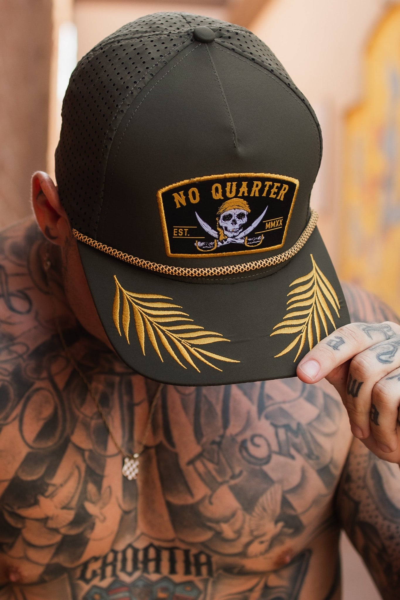 A person wearing the Relentless Betrayals No Quarter Olive Snapback, featuring a skull and crossed swords patch, adorned with yellow palm leaf embroidery on the brim. Tattoos cover their chest and shoulders.