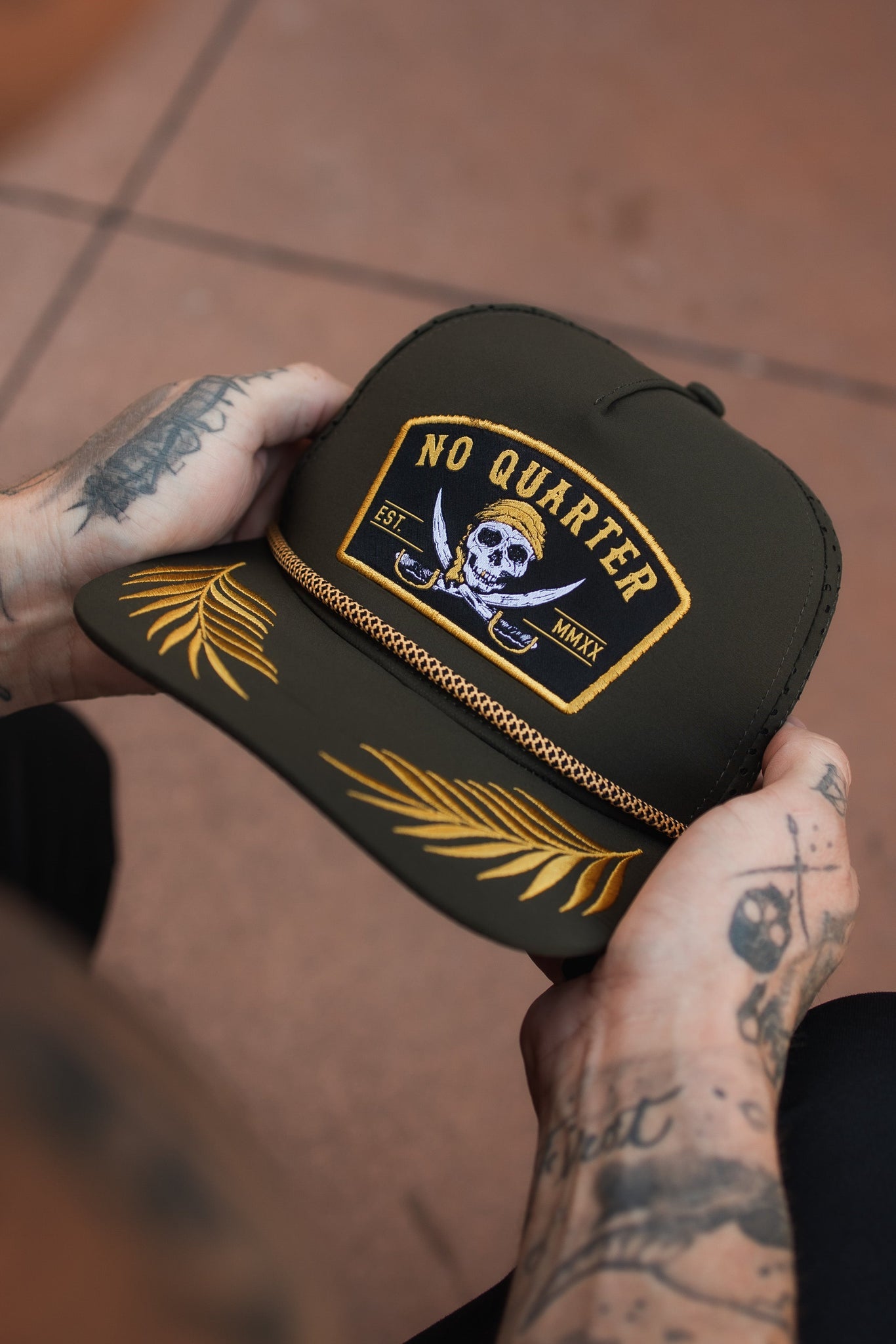 A tattooed individual holds a Relentless Betrayal No Quarter Olive Snapback, showing its skull design, gold embroidered leaves, and braided strap above the brim. The hat is water-resistant and olive in color.