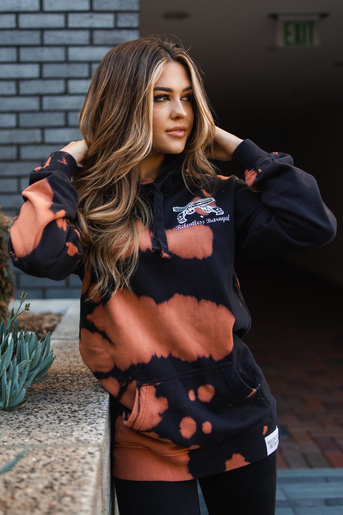 A person with long hair leans against a concrete surface, wearing the Relentless Betrayals No Prey, No Pay Bleach Wash Hoodie in black and orange. Their hands rest on their head as they look to the right. Succulents are in the foreground, with a dimly lit area behind.