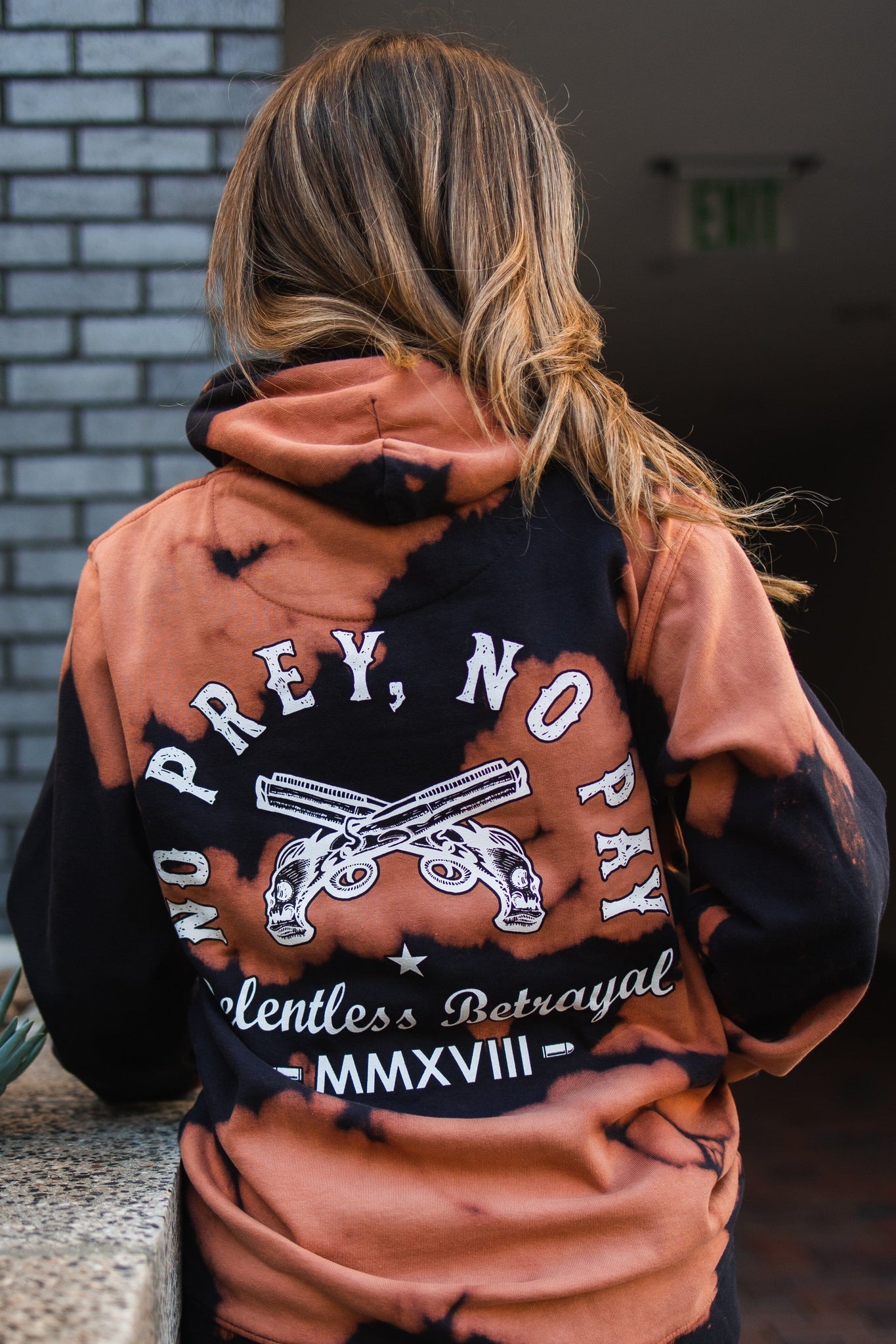 A person with long hair wears a No Prey, No Pay Bleach Wash Hoodie by Relentless Betrayal. The black and orange tie-dye hoodie showcases NO PREY, NO PAY, Relentless Betrayal MMXVIII, and crossed pistols on the back. Made from a cotton-polyester blend fleece, it provides comfort as they turn from the camera.