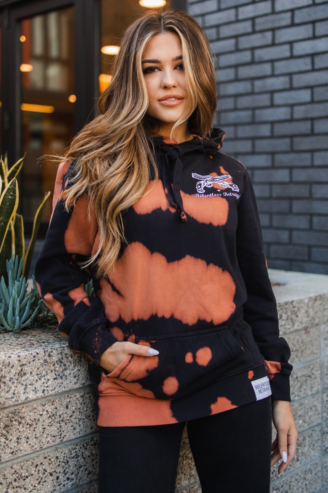 A person with long hair in Relentless Betrayals No Prey, No Pay Bleach Wash Hoodie leans against a brick wall. With one hand in their pocket, they radiate a relaxed vibe. The scene includes lush plants and a building with large windows, enhancing the cozy cotton-polyester blend fleece of the hoodie.