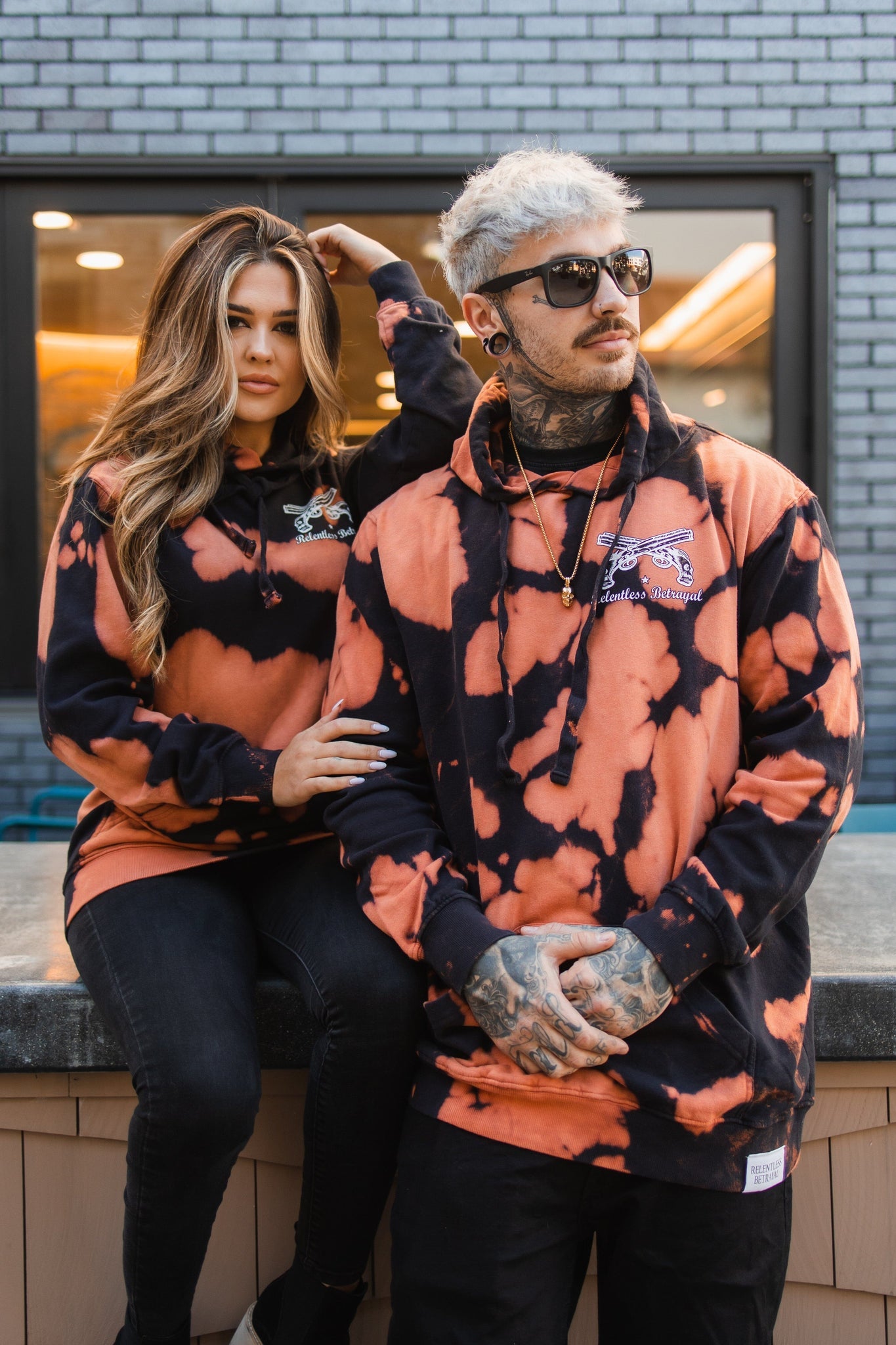 Two tattooed individuals confidently pose in matching orange and black No Prey, No Pay Bleach Wash Hoodies from Relentless Betrayal. One sports stylish sunglasses and stands with crossed arms, while the other sits with their hand on the others shoulder. Both rock these unisex cotton polyester blend fleece hoodies.
