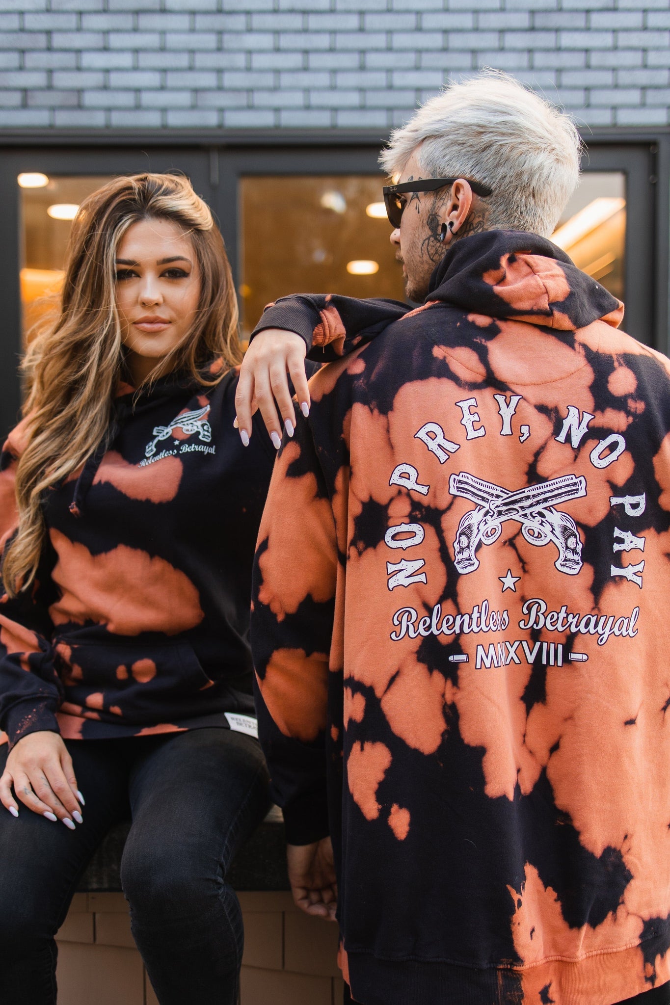 A woman sits on a ledge wearing a Relentless Betrayal black and orange bleach tie-dye hoodie with a front logo. Next to her, a man stands in the unisex No Prey, No Pay version featuring pistols and designs on the back. Both hoodies are made from cotton polyester blend fleece for comfort and style.