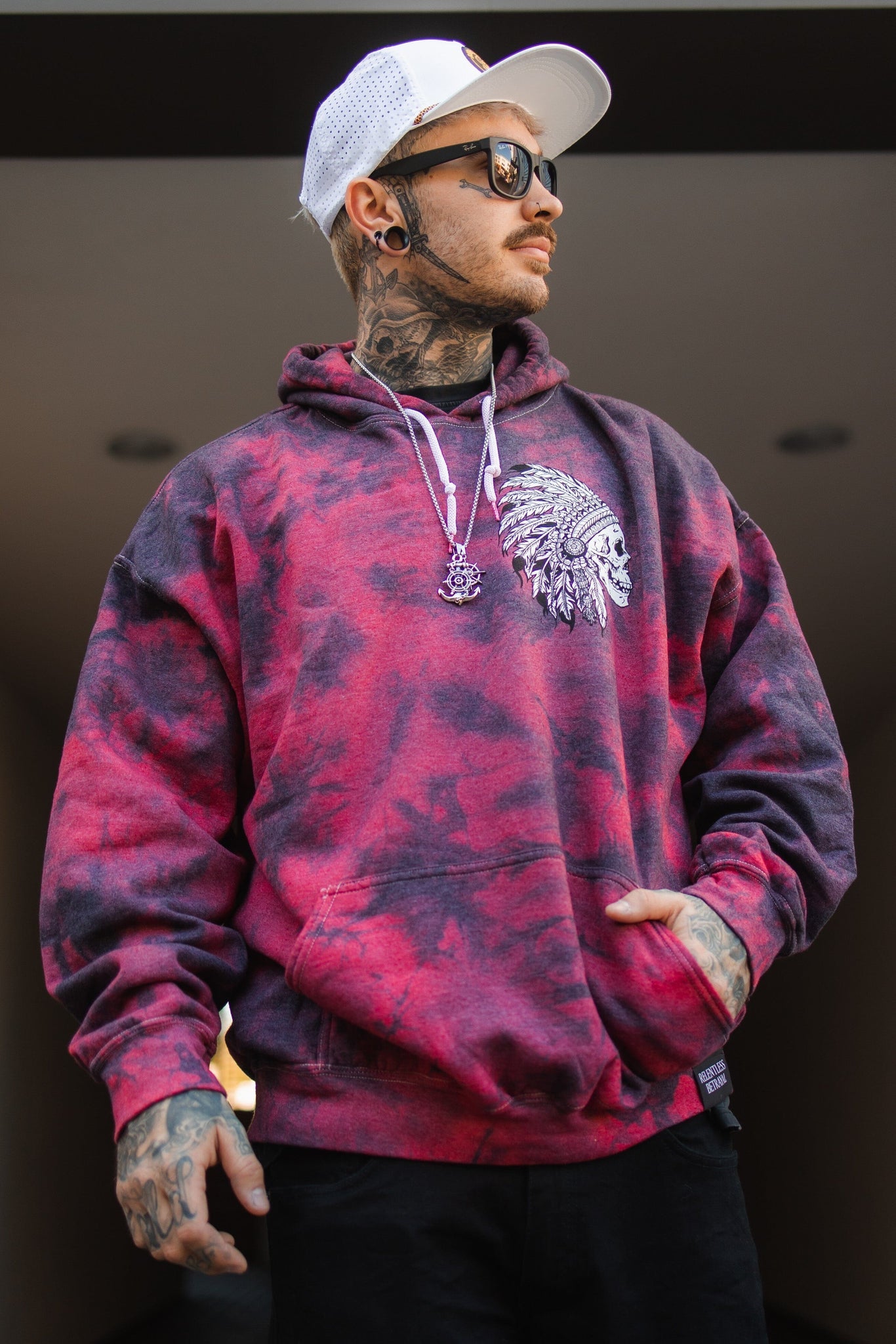 The man wears a Relentless Betrayal Trustless Chief Red Tie Dye unisex hoodie featuring a Native American chief graphic. Paired with sunglasses, a white cap, and black pants, his tattoos are on display as he gazes to the side with one hand casually in his pocket.