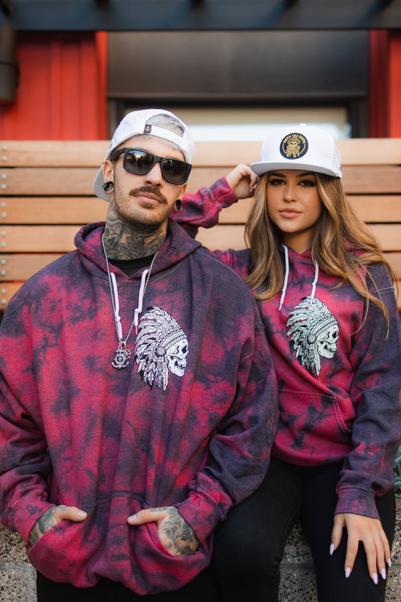 A man and woman sport matching Trustless Chief Red Tie Dye Hoodies by Relentless Betrayal, showcasing a Native American chief design. Both wear white caps; the woman sits on a bench as the man stands beside her, their cotton/polyester blend adding casual flair to their outdoor vibe.