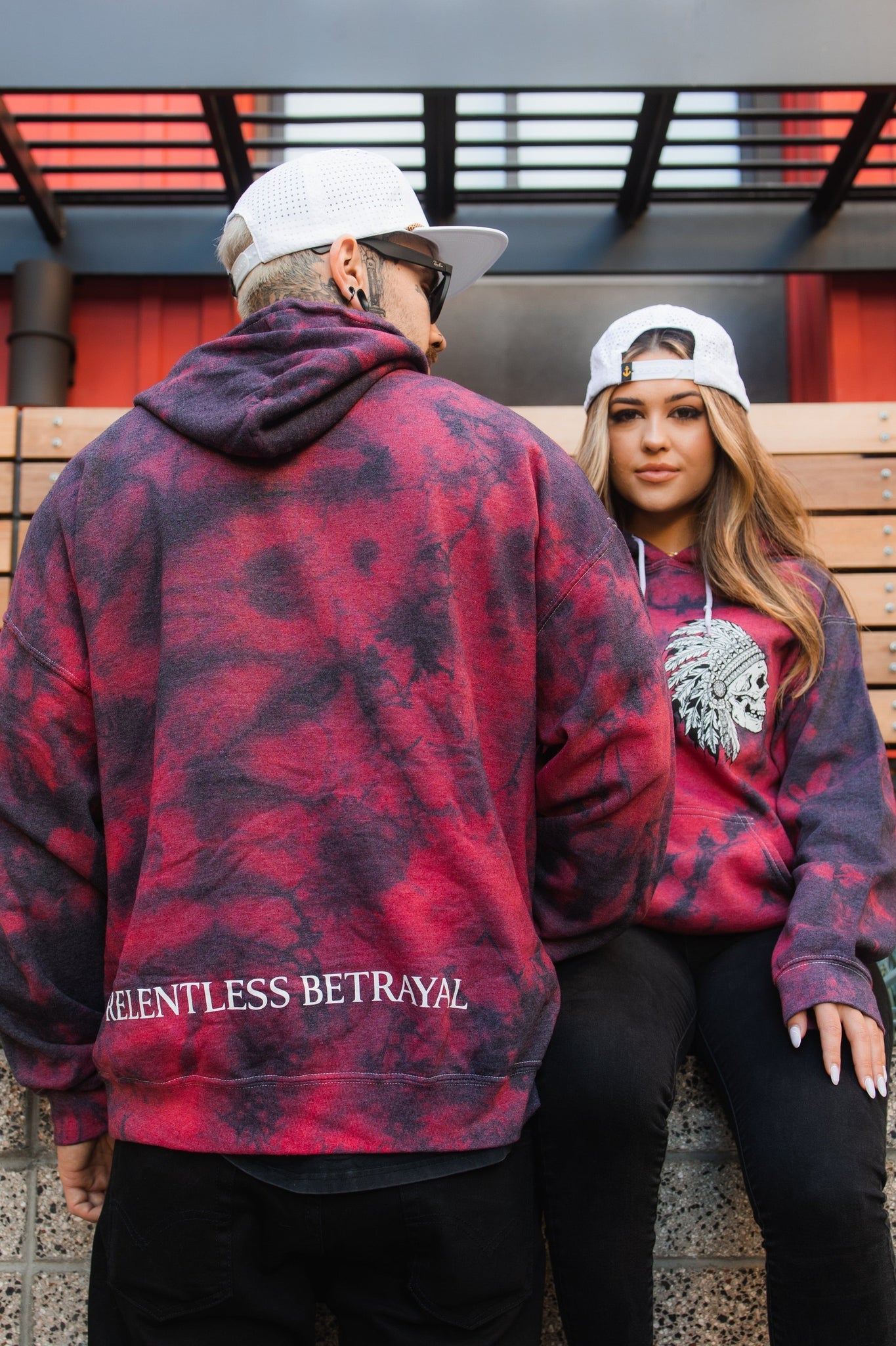 Two people in matching Trustless Chief Red Tie Dye Hoodies sit outside. The person on the left has RELENTLESS BETRAYAL on the back of their unisex hoodie. Both wear caps and sunglasses, with one sitting on a bench and the other leaning casually against it.