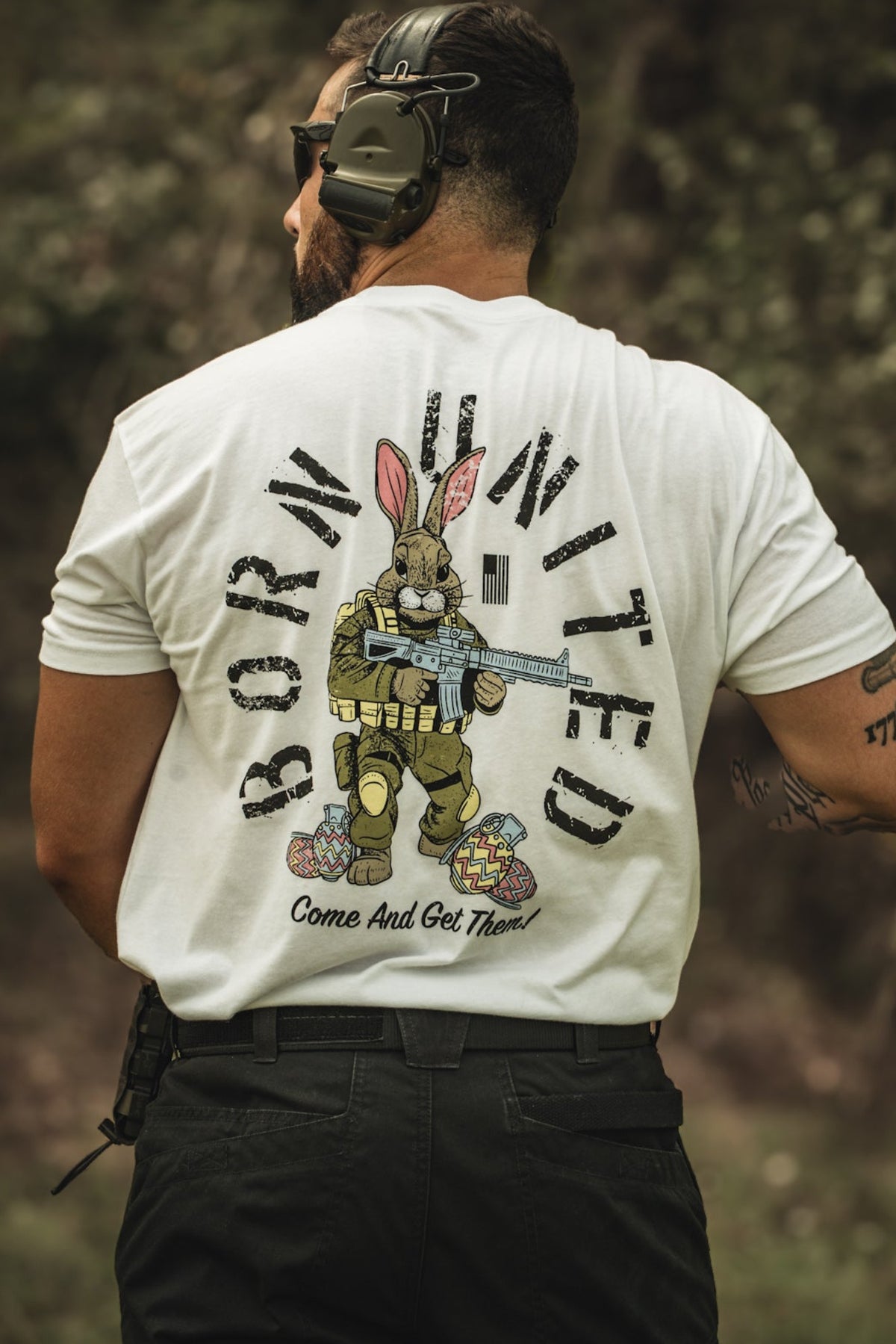 A man wearing hearing protection and a Born United Private Label "Battle Bunny" T-shirt, featuring a cartoon bunny in military gear with "BORN UNITED" and "Come And Get Them," stands outdoors facing away from the camera.