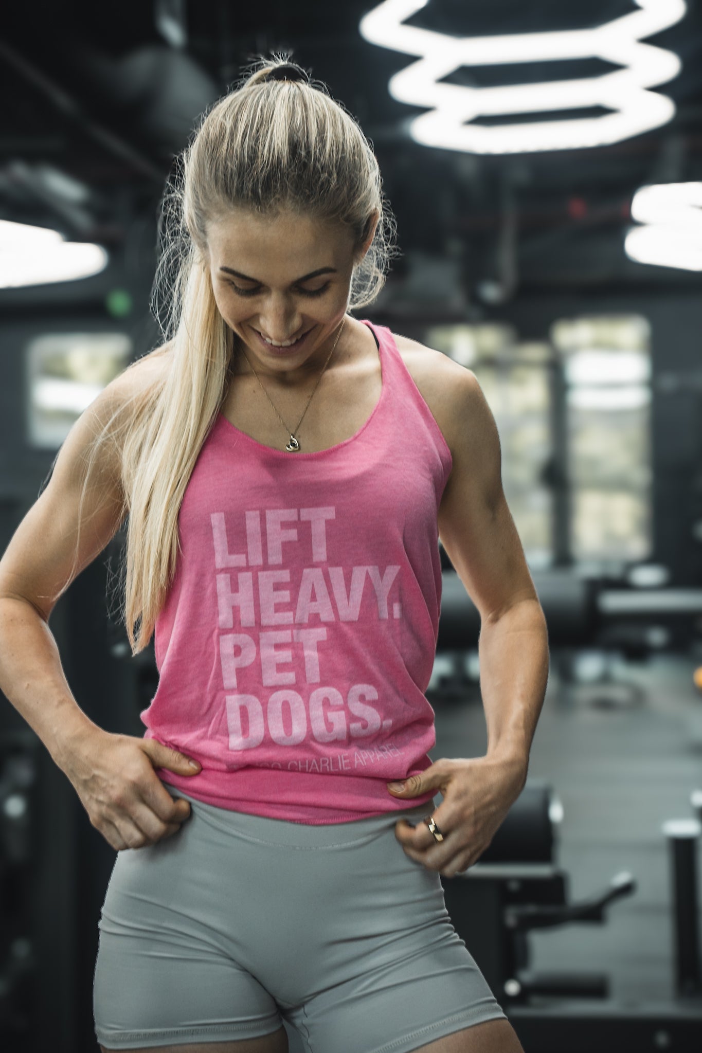 A woman with long blonde hair smiles and looks down at her Tango Charlie Apparel Lift Heavy. Pet Dogs. Womens Pink Tank, standing in a gym in light gray shorts with workout equipment behind her.