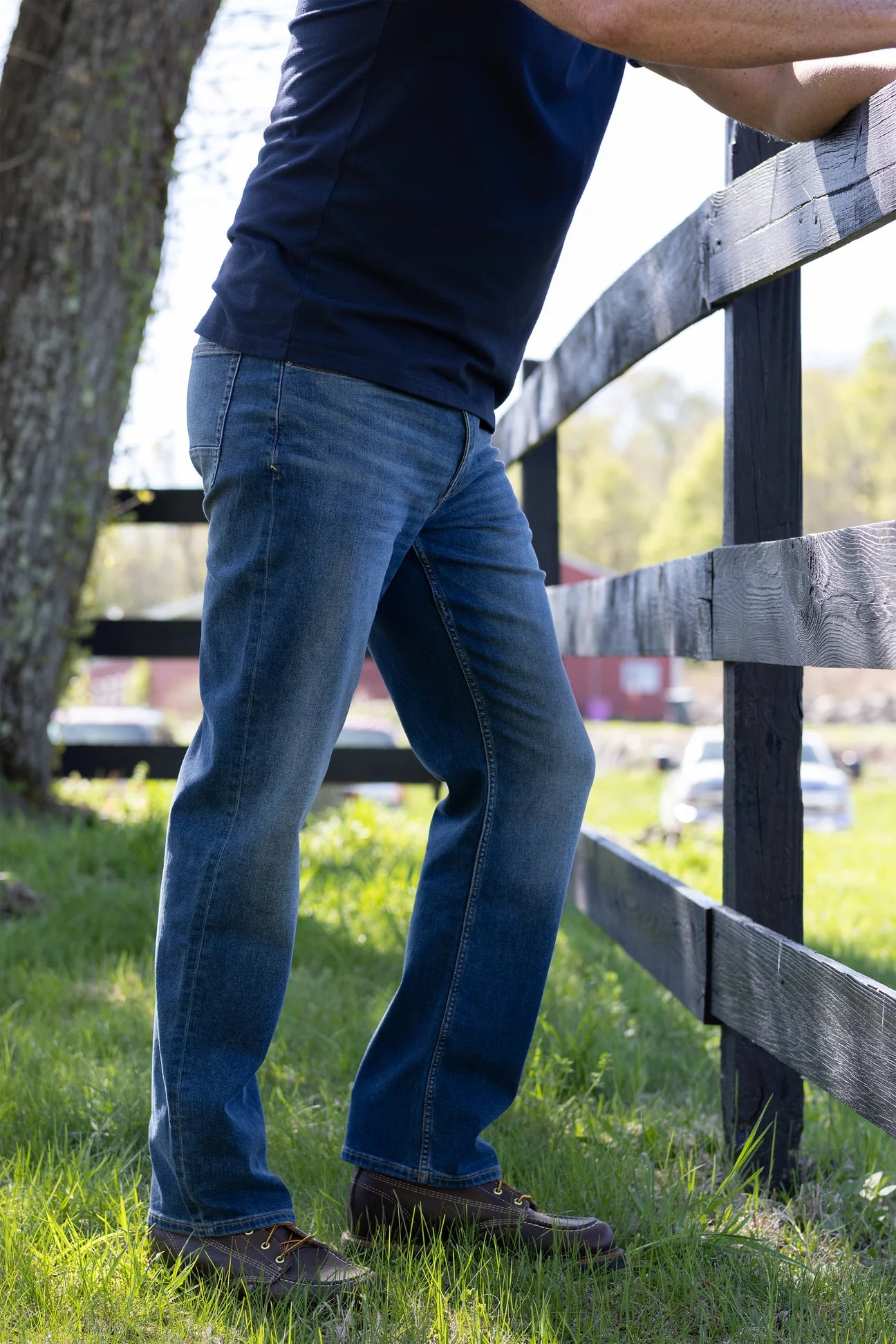 Wearing Devil Dog Dungarees Relaxed Straight Jean - Bryson City, a person in a navy shirt and brown shoes stands on grass by a wooden fence in sunlight.