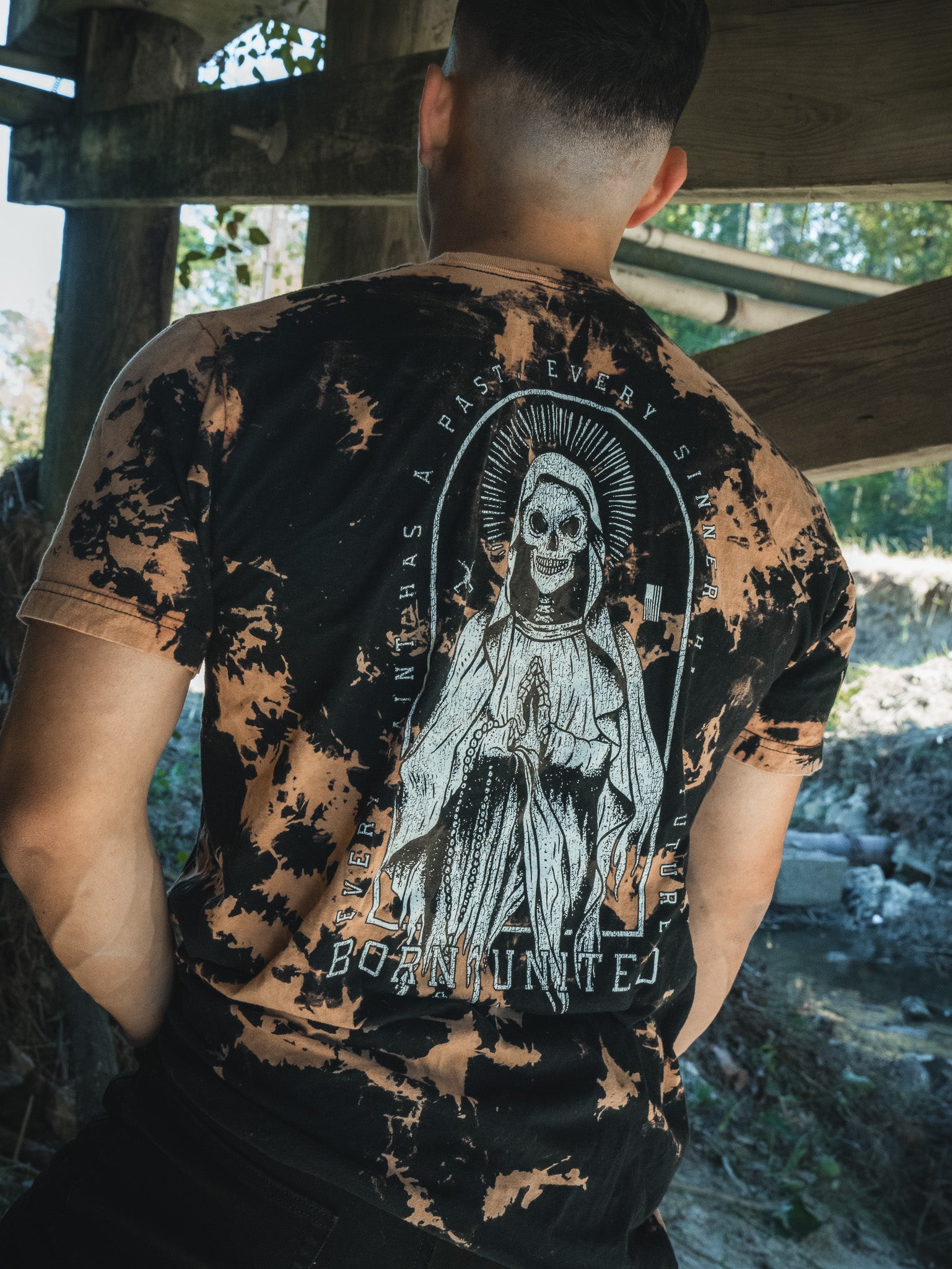 A person stands outdoors under a wooden structure, wearing the Saints & Sinners- Bleach Wash tee by Born United Private Label. The back features a detailed skeleton with a halo, draped in robes, surrounded by redemption-themed text. Trees and a stream evoke hardcore serenity in the background.