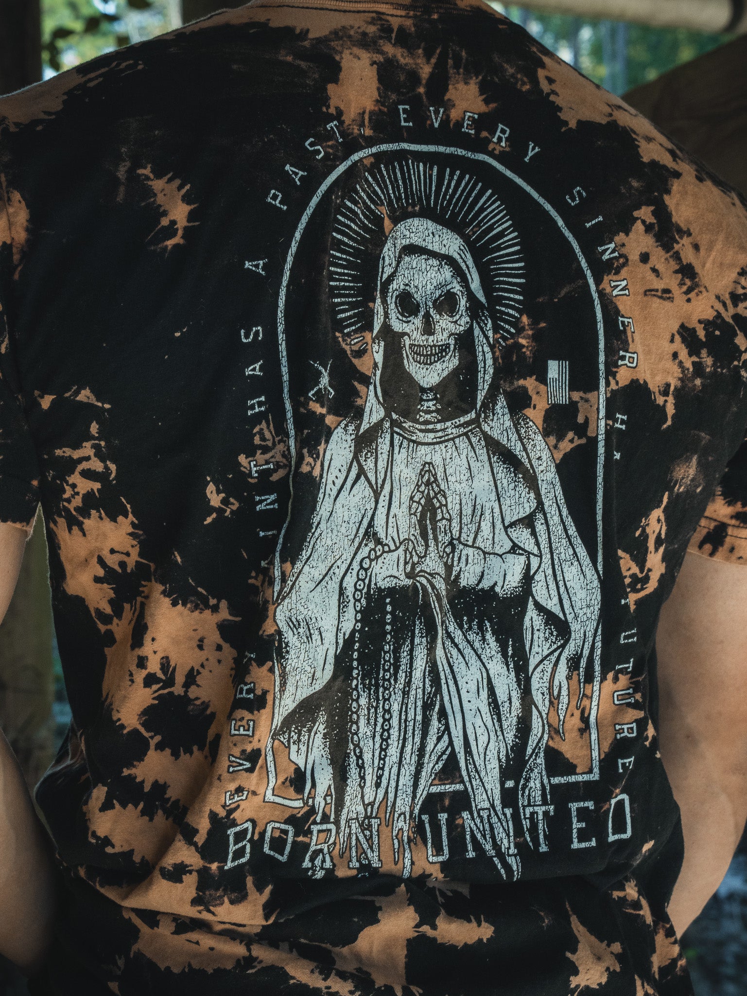A person wears the Saints & Sinners-Bleach Wash tee by Born United Private Label. This black and brown shirt features a graphic of a skeleton in a robe with clasped hands, surrounded by the words EVERY SAINT HAS A PAST EVERY SINNER HAS A FUTURE and BORN UNITED, symbolizing redemption.