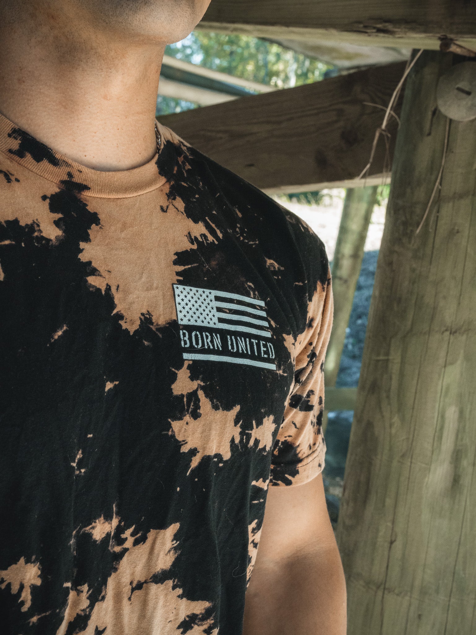 A person wearing the Saints & Sinners- Bleach Wash tee by Born United Private Label, featuring an American flag patch and Born United, is partly visible against a backdrop of wooden beams and greenery, creating a hardcore vibe.