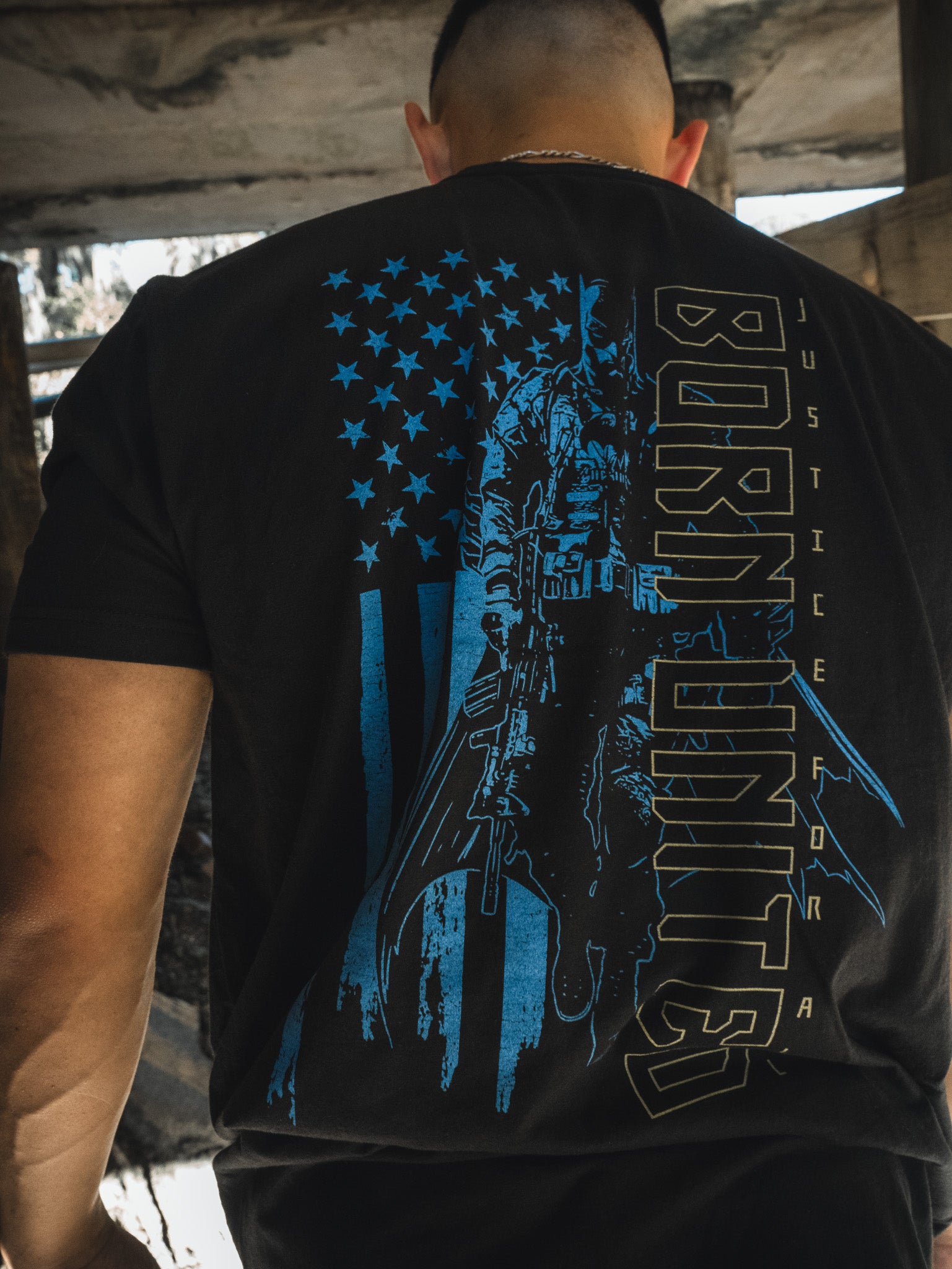 With his back to the camera, a man wears a Born United Private Label Justice For All tee. The black shirt prominently features a blue and black American flag design, an iconic figure with bat-like wings holding a weapon, and the phrases BORN UNITED and JUSTICE FOR ALL.