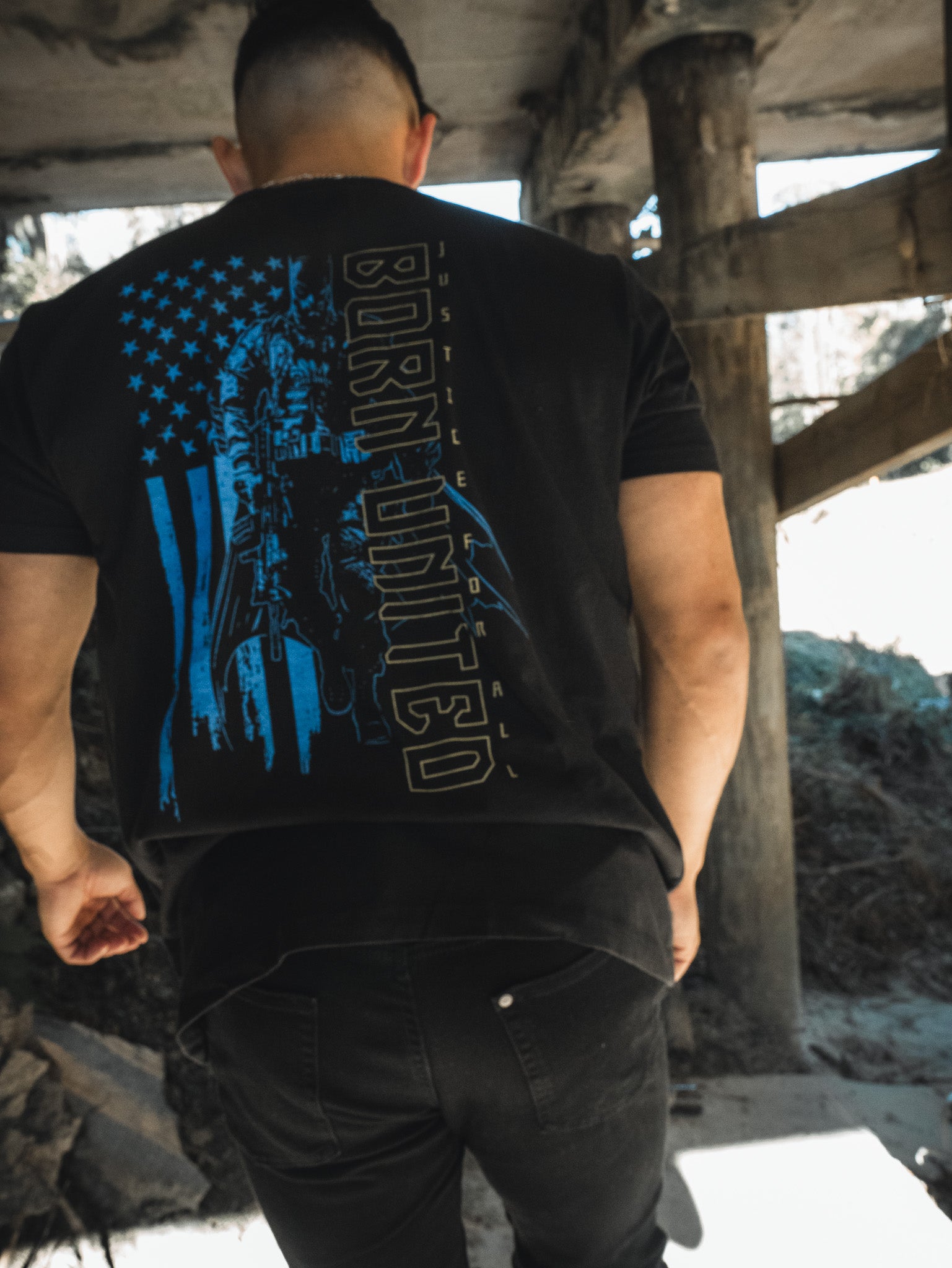 A person with short hair wears a Justice For All tee from Born United Private Label, featuring a blue design and BORN UNITED, as they stroll through a rugged, Gotham-like outdoor setting with sunlight filtering through a structure.