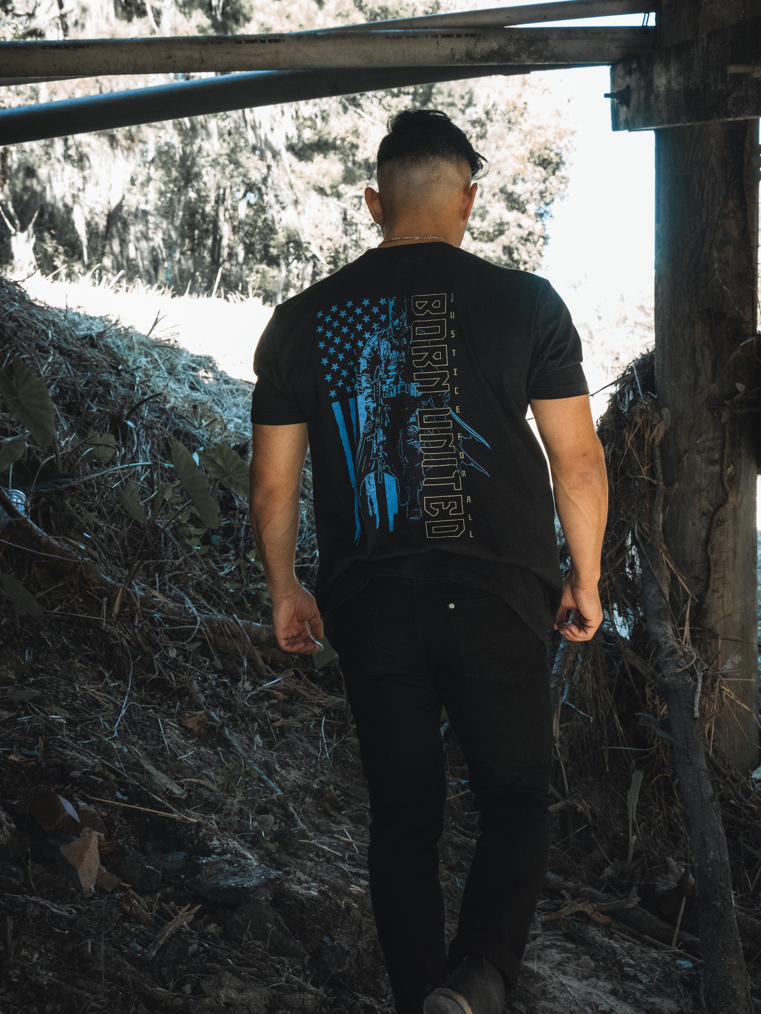 A person with short hair wearing Born United Private Labels Justice For All original printed tee strolls under a structure along a natural path, surrounded by foliage. With their back to the camera, they explore the outdoors like one of Gothams masked heroes in an urban jungle.