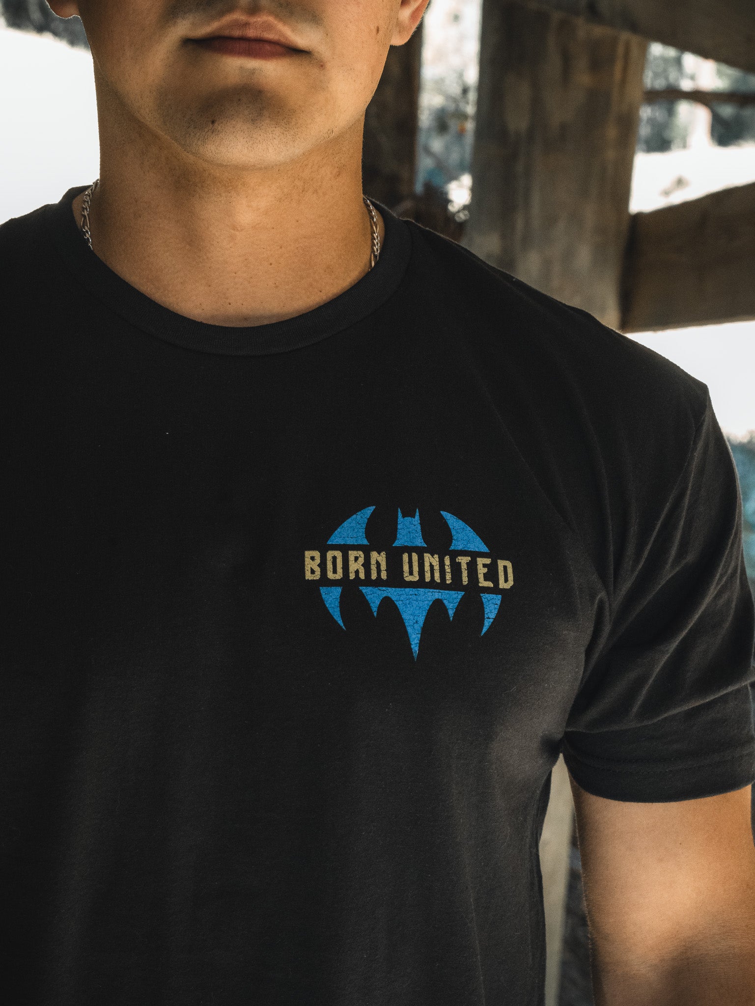 Partially shadowed, a person wears the Justice For All tee from Born United Private Label. The original design showcases a blue bat logo and Born United, evoking the spirit of Gothams masked heroes.