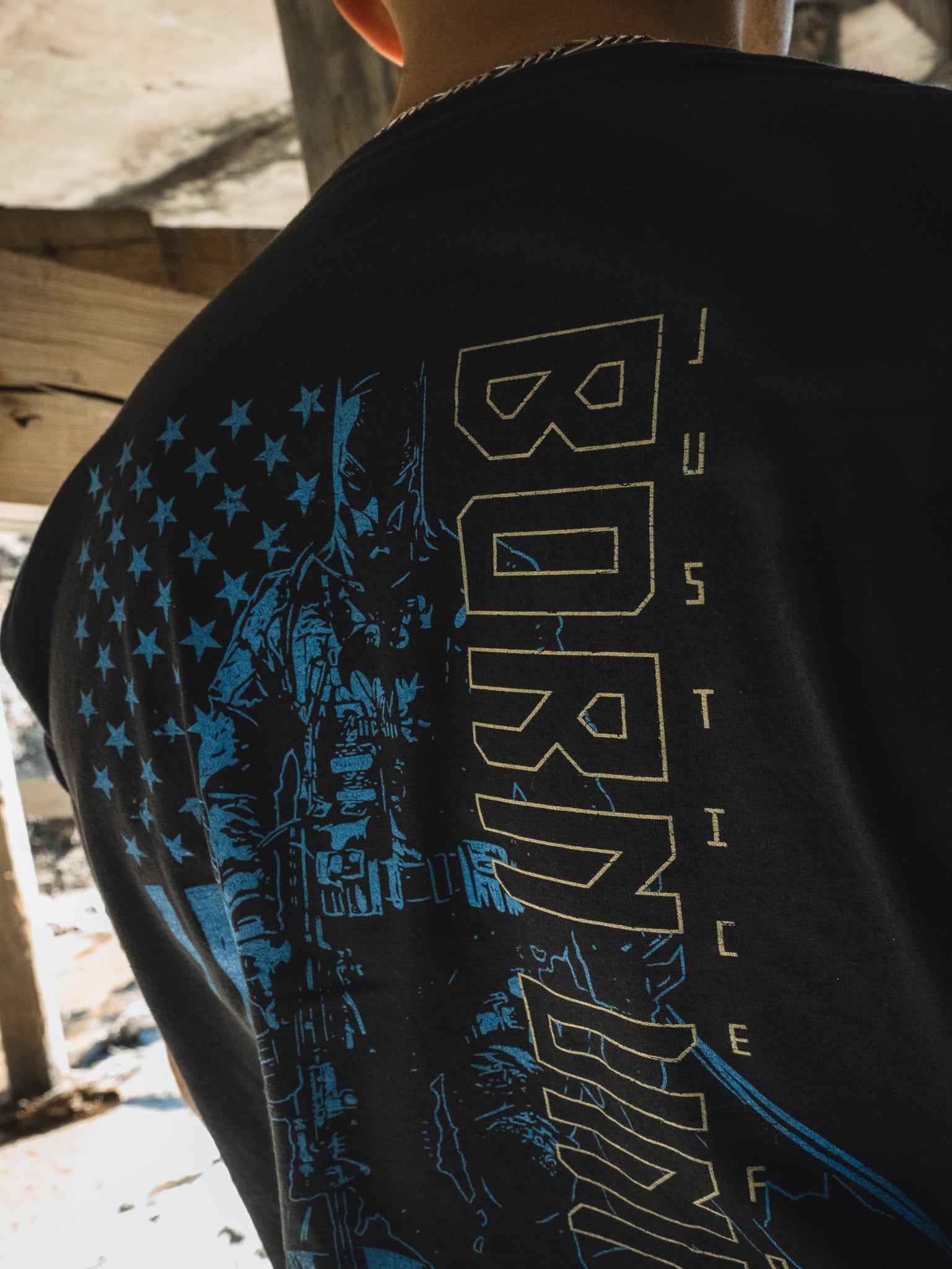 A person wearing the Justice For All tee from Born United Private Label, featuring an American flag, soldier illustration, and the words BORN and JUSTICE, evokes the vibe of masked heroes in Gotham.