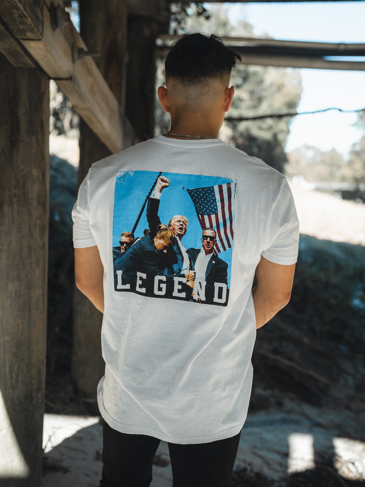 A person models the Forever Legend - Trump t-shirt by Born United Private Label, showing off its premium white fabric with a people-and-flag graphic and the text Forever Legend. The serene outdoor backdrop includes wooden structures and greenery.