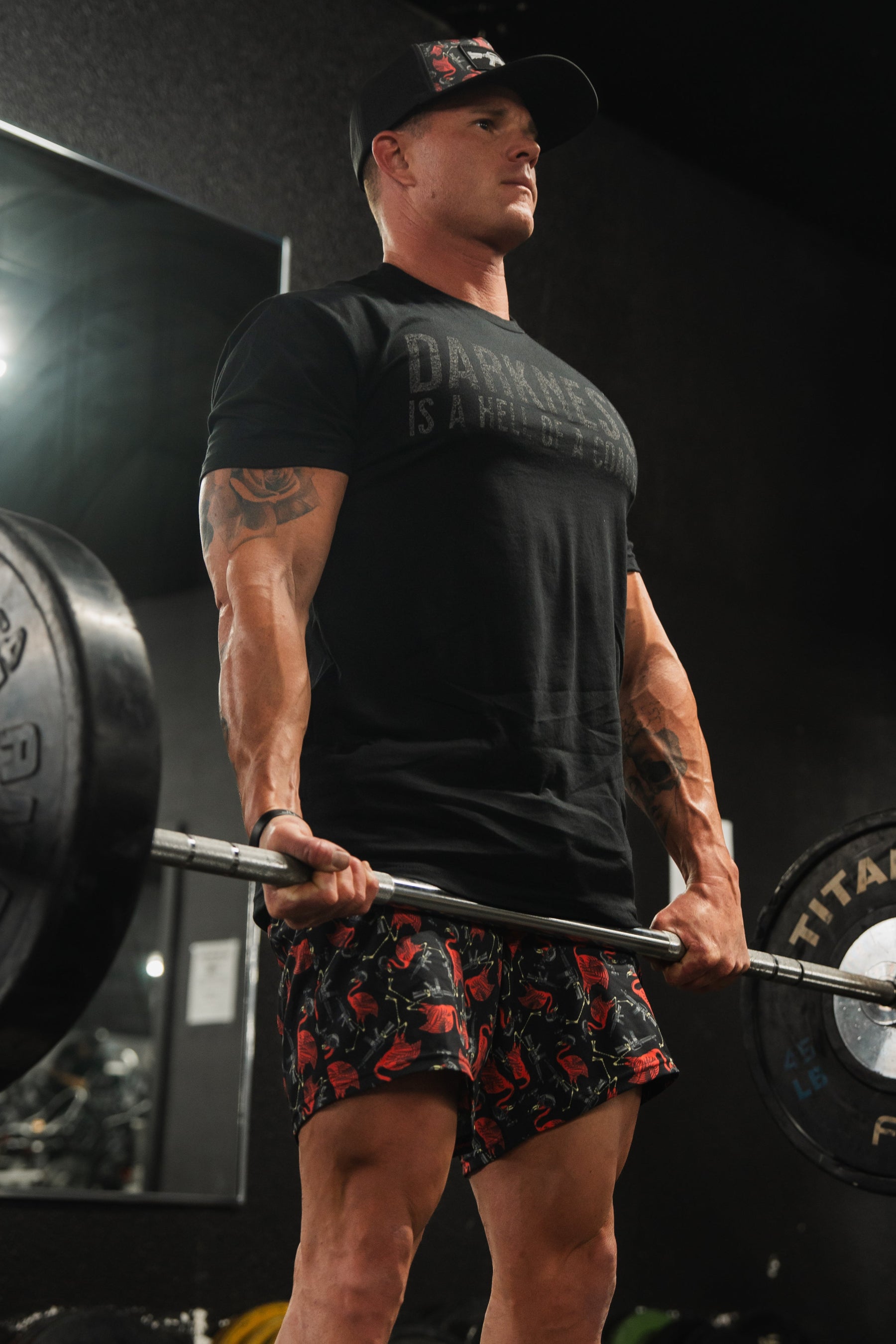 In a gym with dark walls and a reflective mirror, someone in a black cap and T-shirt is deadlifting while wearing Tango Charlie Apparels Mens Performance Training Shorts 5.5 - Tactimingo, gripping a barbell loaded with weights.