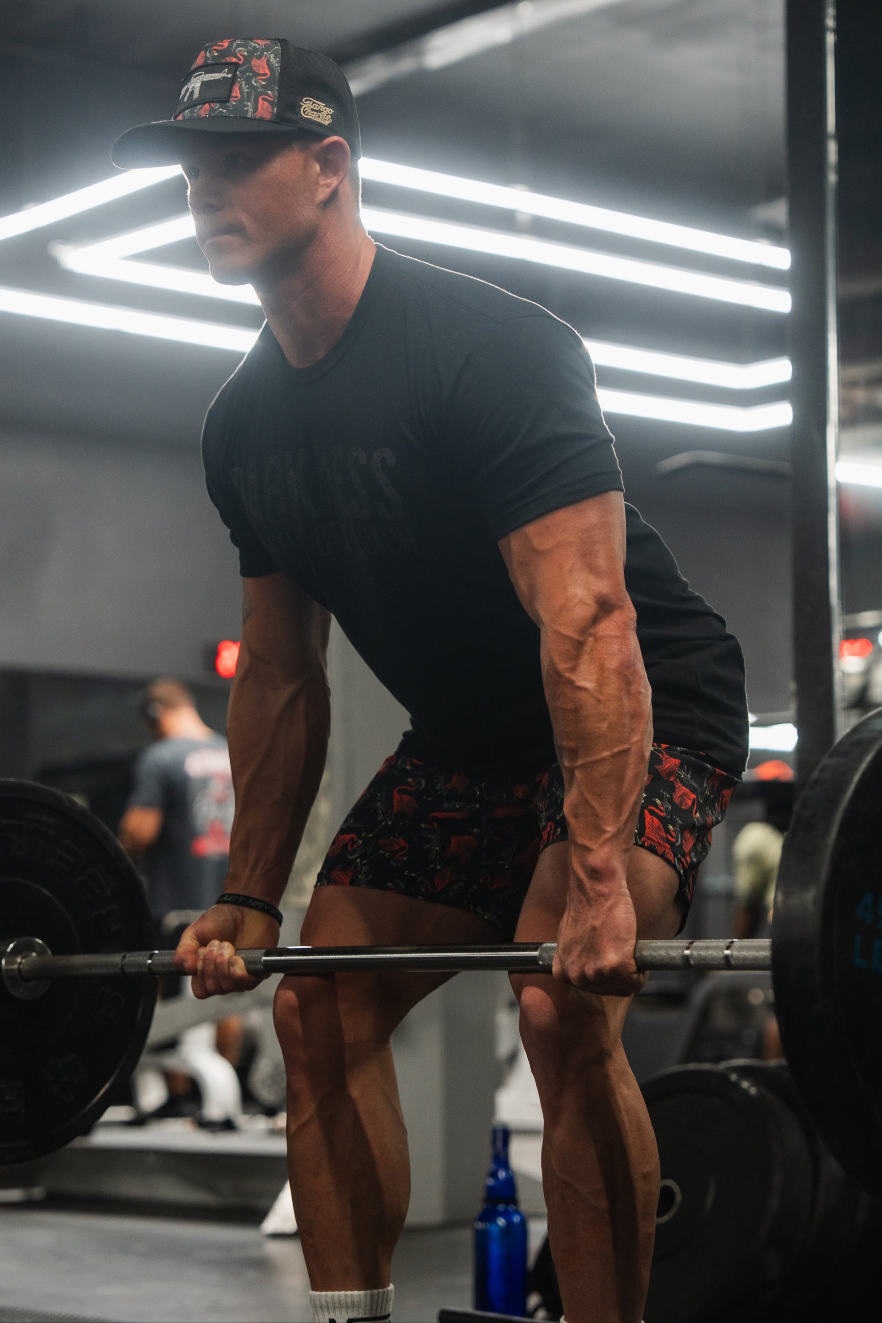 In a dimly lit gym, a person in a black T-shirt and Tango Charlie Apparels Mens Performance Training Shorts | 5.5- Tactimingo lifts a barbell with focused determination. Bright ceiling lights illuminate the scene, while versatile gym equipment stands ready in the background.