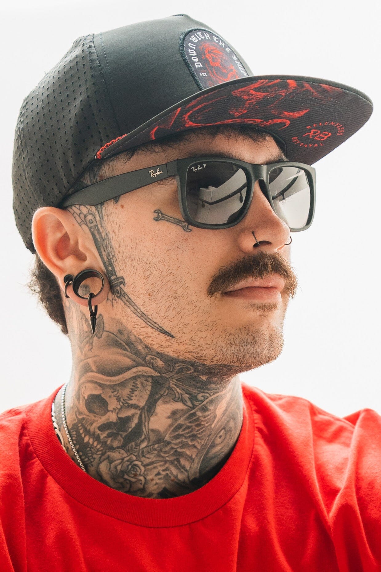 A man wears Relentless Betrayals Ship Wreck Snapback - Red, paired with a red shirt. He sports tattoos on his neck, a mustache, sunglasses, a nose ring, ear gauges, and a black cap with its striking design. His expression is neutral.