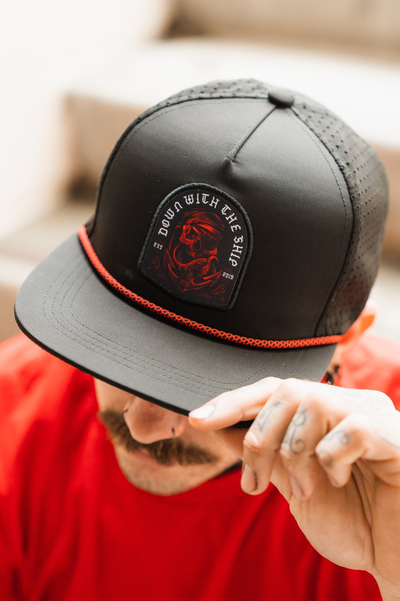 A man in a red shirt adjusts his Ship Wreck Snapback - Red from Relentless Betrayal. The high-quality cap, adorned with a skull and the phrase DOWN WITH THE SHIP, features a logo and red trim. His tattooed hand ensures its perfectly fitted for any occasion.