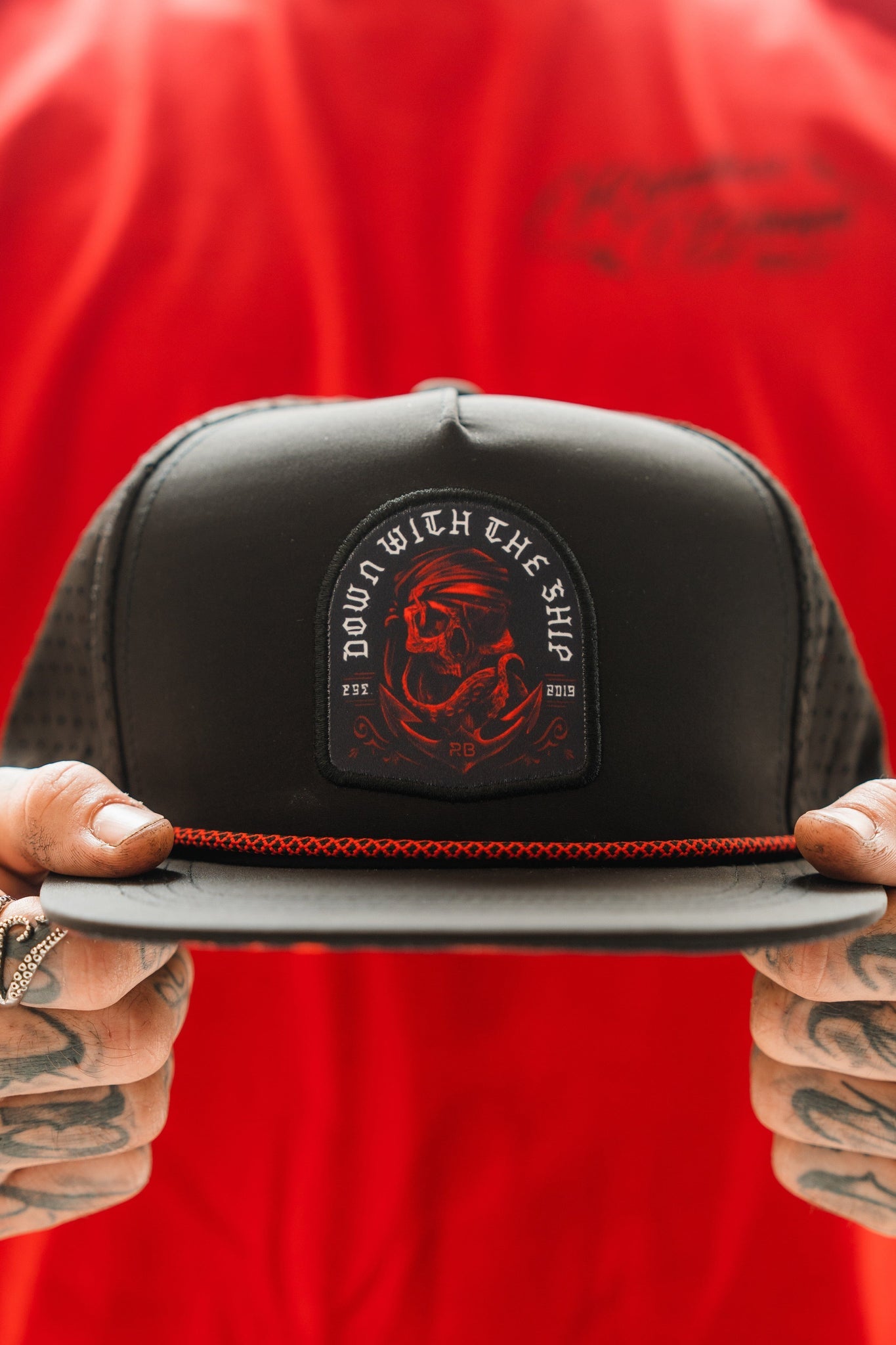 A person sports the Relentless Betrayal Ship Wreck Snapback - Red, boasting a skull and crossbones patch with DOWN WITH THE SHIP text. This water-resistant accessory pairs perfectly with their red shirt, rings, and tattooed fingers for an edgy style.