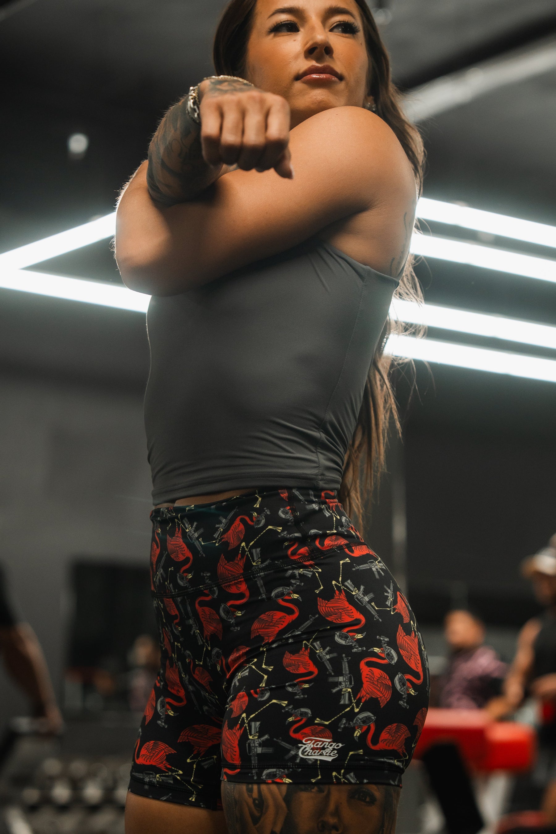 In a gym with bright lights and blurred figures, a person stretches in workout attire wearing Tango Charlie Apparels gray top and Womens High-Waisted Seamless Front Compression Training Shorts by Tactimingo. The breathable fabric ensures comfort as they raise their arm overhead.