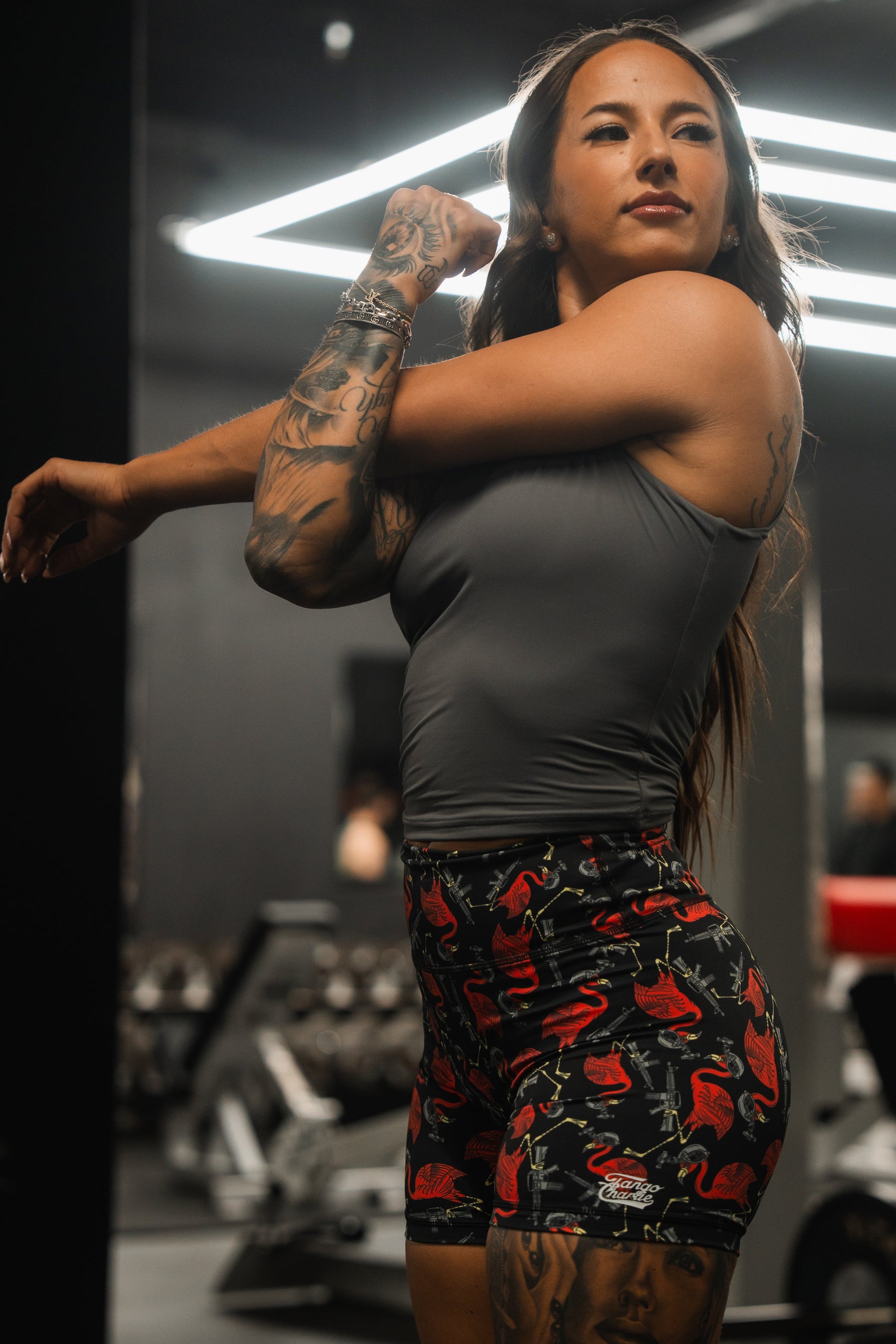 A woman stretches her arm across her chest at a gym with modern equipment, wearing a gray sleeveless top and vibrant Womens High-Waisted Seamless Front Compression Training Shorts by Tango Charlie Apparel. Her arms are tattooed, complementing the breathable fabric of her stylish outfit.