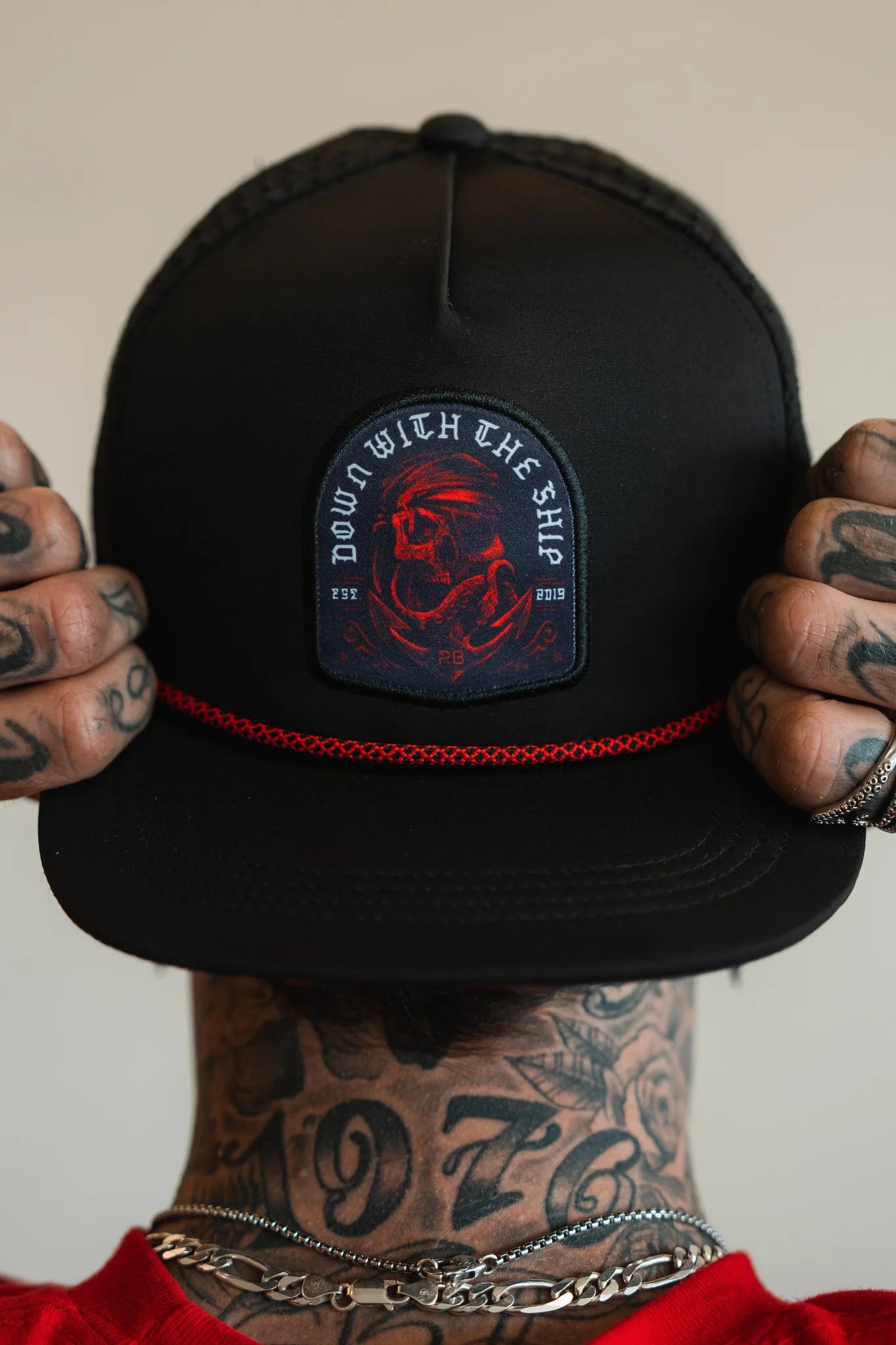 A person holds the brim of a Relentless Betrayal Ship Wreck Snapback, revealing tattoos on their fingers and neck. This black cap features a round patch with a red art design and DOWN WITH THE SHIP phrase. They accessorize with silver rings and a necklace.