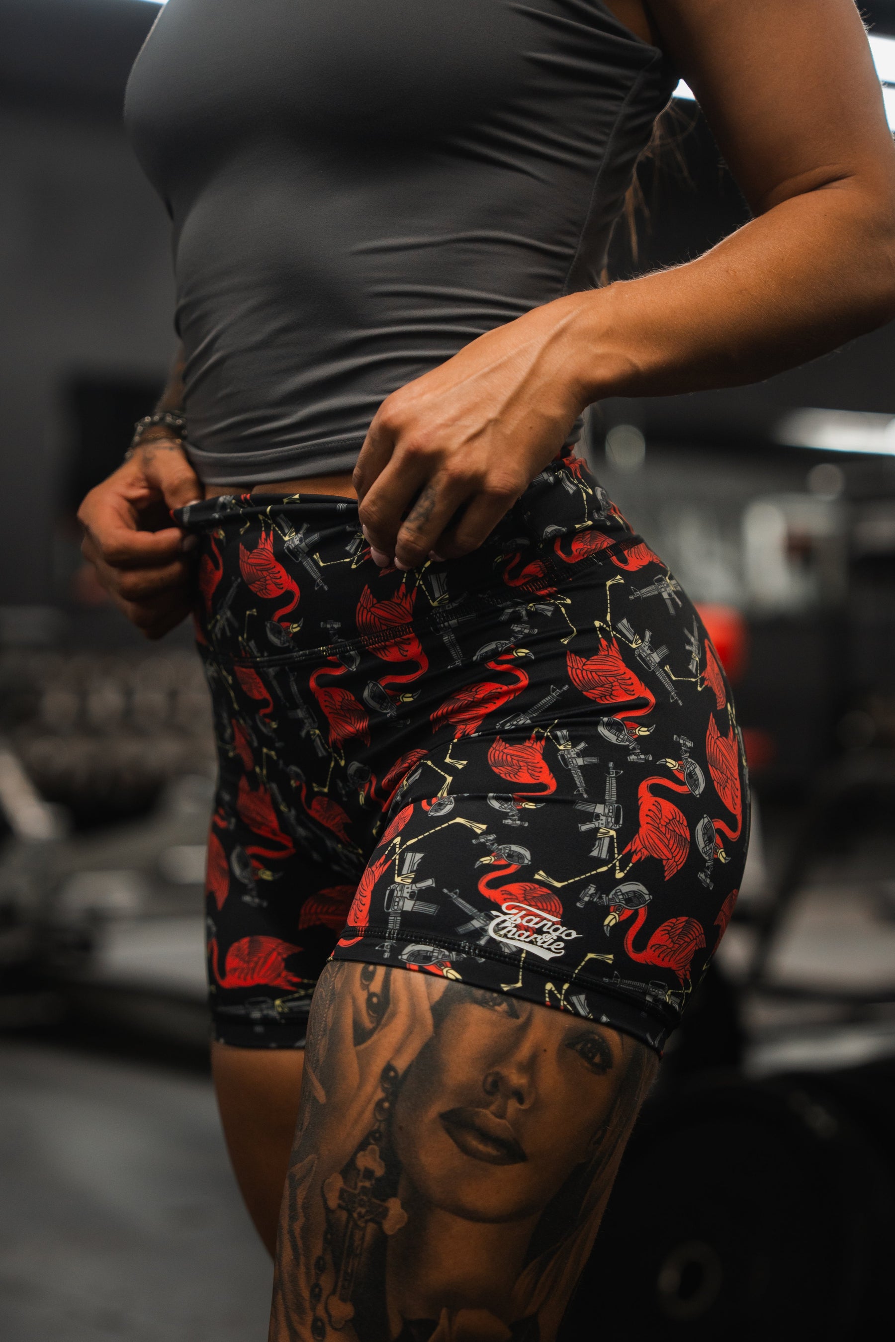 At the gym, someone sports Tango Charlie Apparels Tactimingo gray tank top with Womens High-Waisted Seamless Front Compression Training Shorts, adorned with red flamingos and music designs. Their tattooed arm stands out, while the breathable fabric ensures workout comfort.